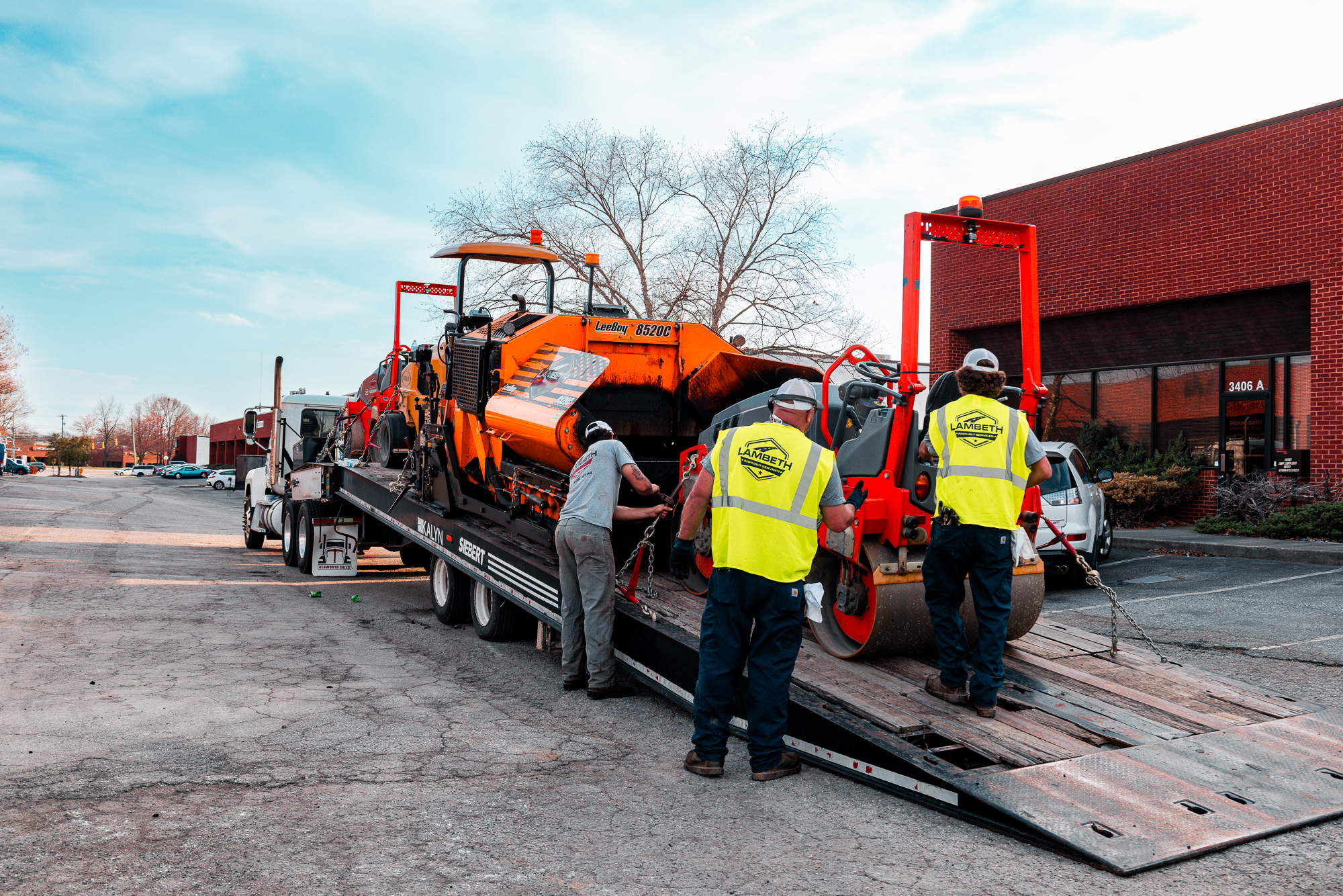 Three workers in lambeth high-visibility vests securing construction machinery on a flatbed truck in a parking lot.