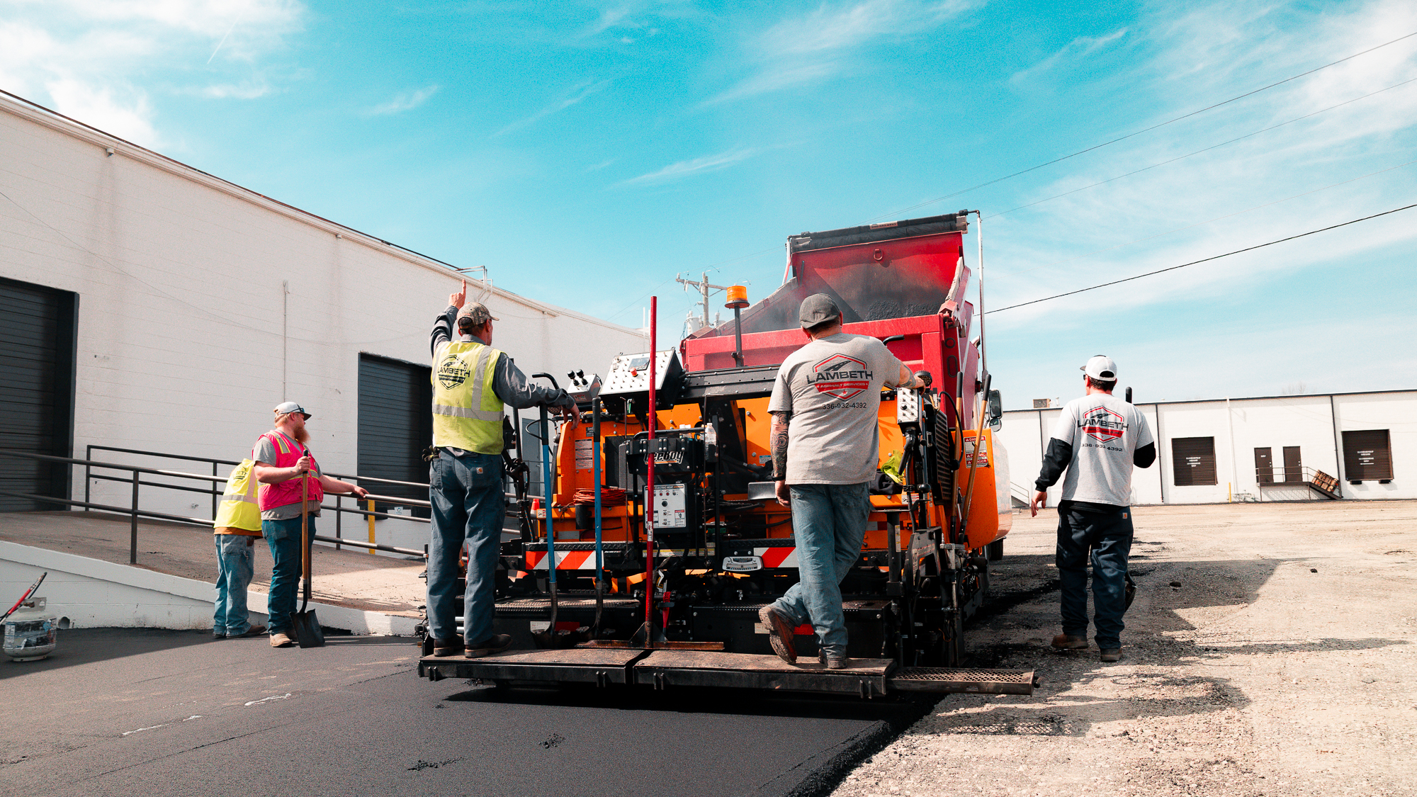 Workers in safety vests and workwear operating machinery to pave asphalt on an industrial lot next to white buildings under a blue sky.