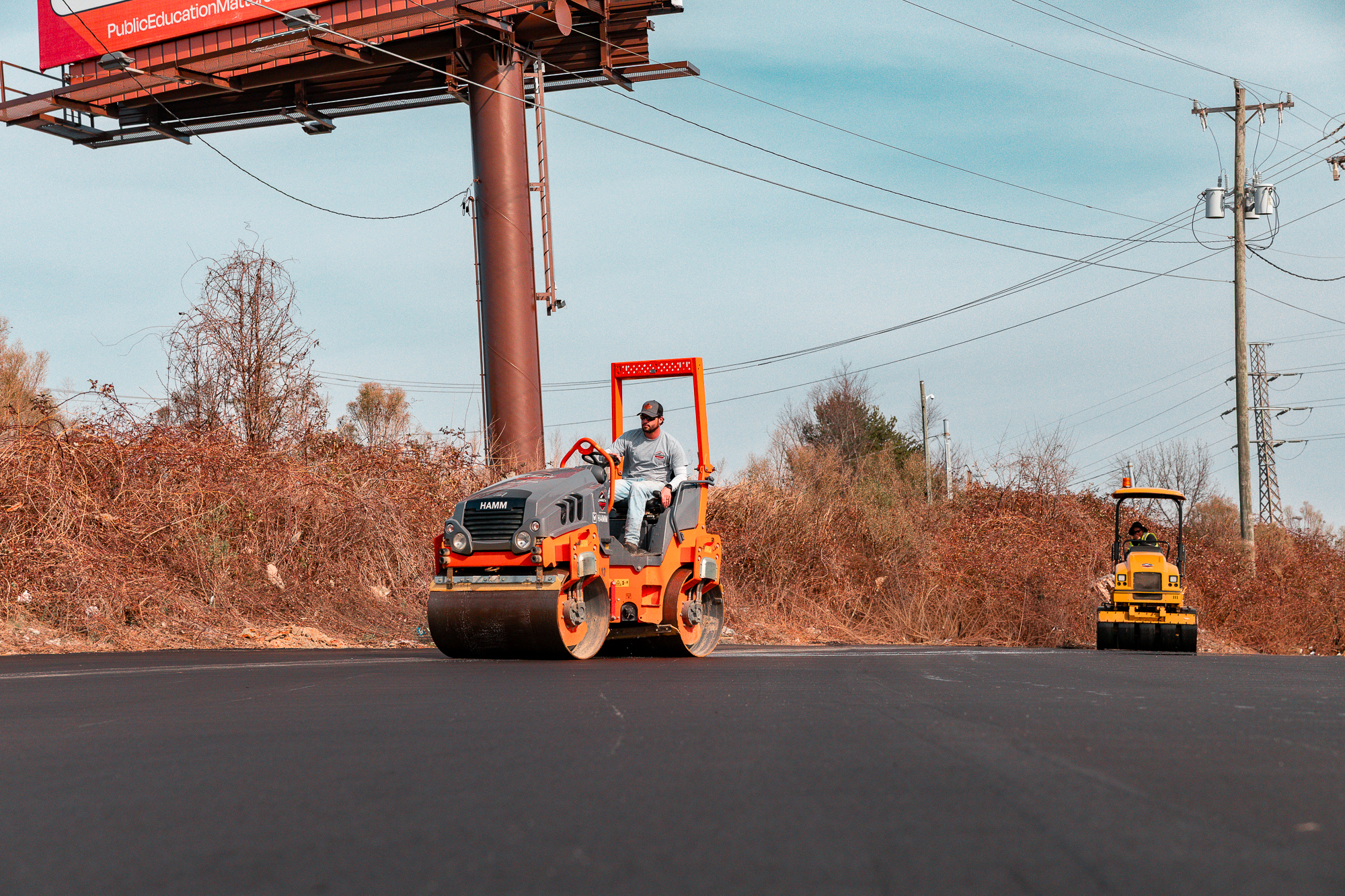Two construction workers operating road rollers on freshly paved asphalt under a blue sky near dry vegetation and power lines.
