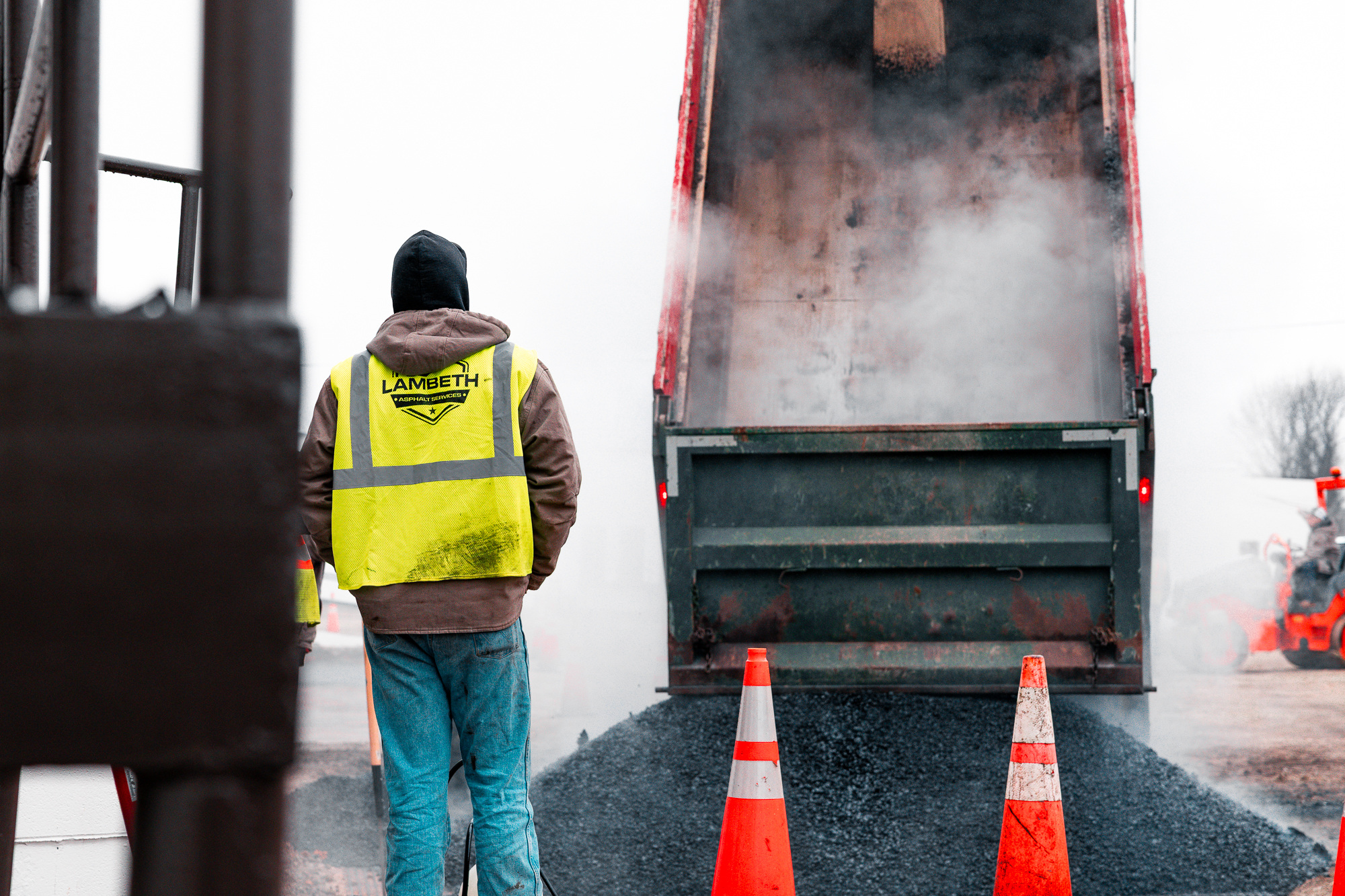 Construction worker facing away wearing a Lambeth Asphalt Services safety vest standing near a truck unloading hot asphalt.