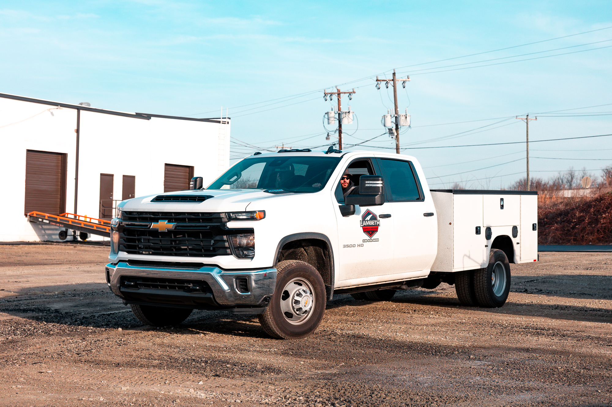 White Chevrolet 3500 HD utility truck with company logo parked on gravel lot near white building.