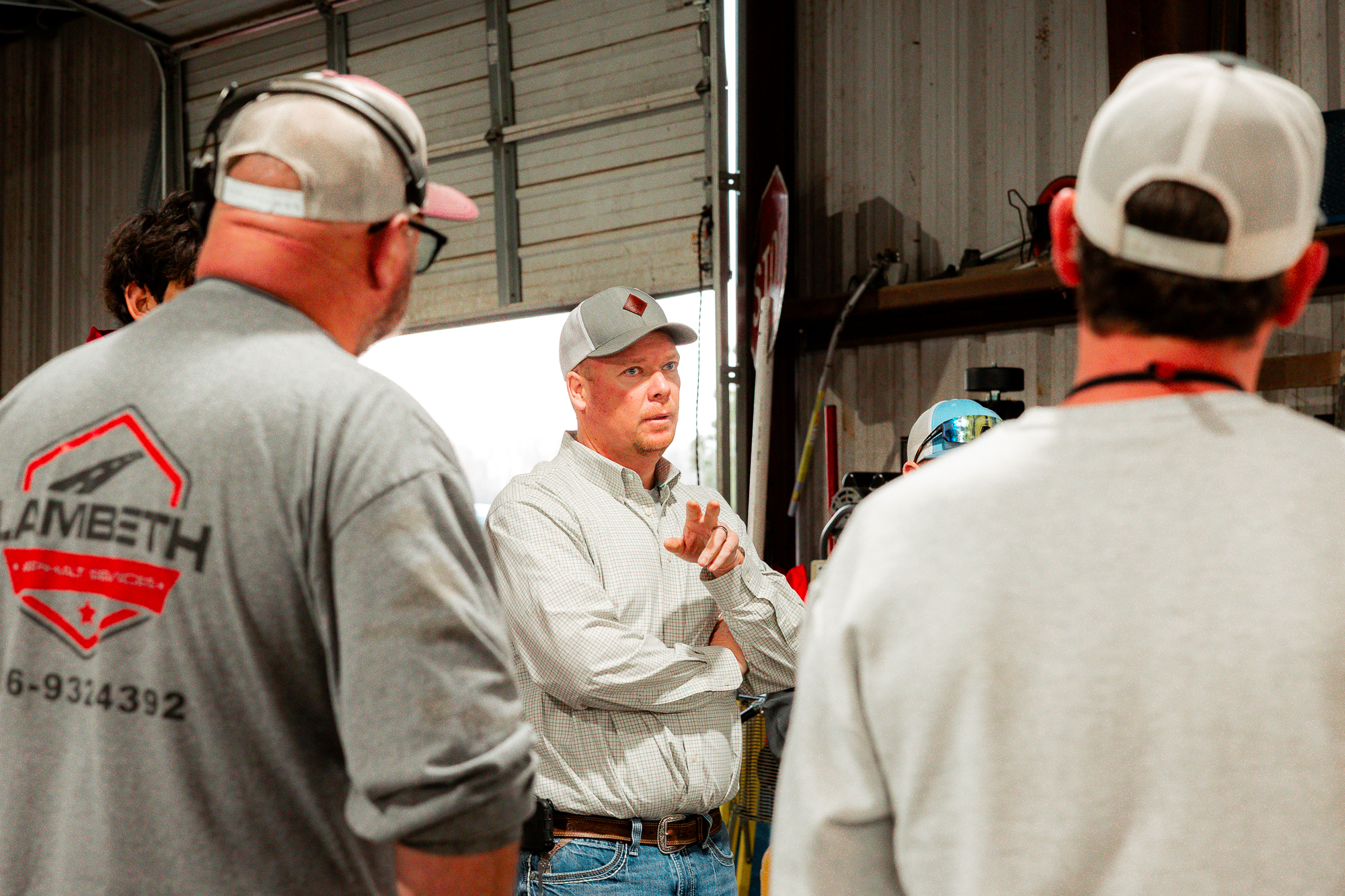 Man in a checkered shirt and baseball cap speaking and gesturing to a small group inside a workshop.