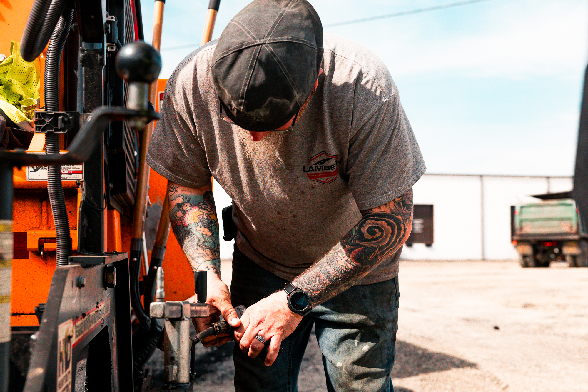 Tattooed man wearing a gray shirt and cap working on machinery outdoors.