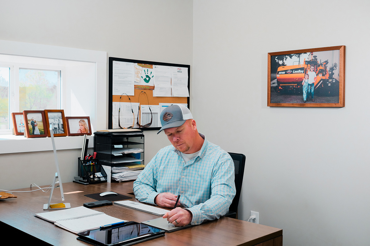 Man in a plaid shirt and cap using a digital pen on a tablet at a desk with a desktop computer and family photos in the background.
