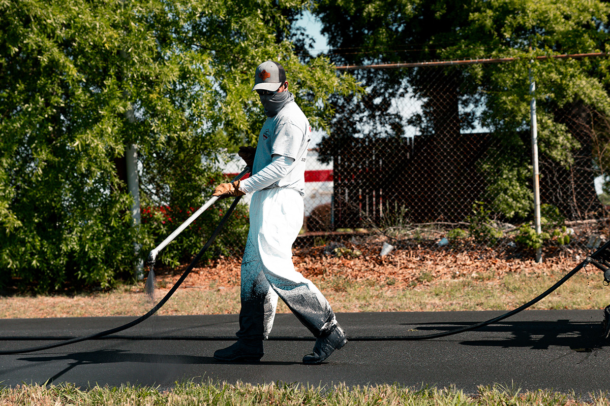 Workers in safety vests and workwear operating machinery to pave asphalt on an industrial lot next to white buildings under a blue sky.
