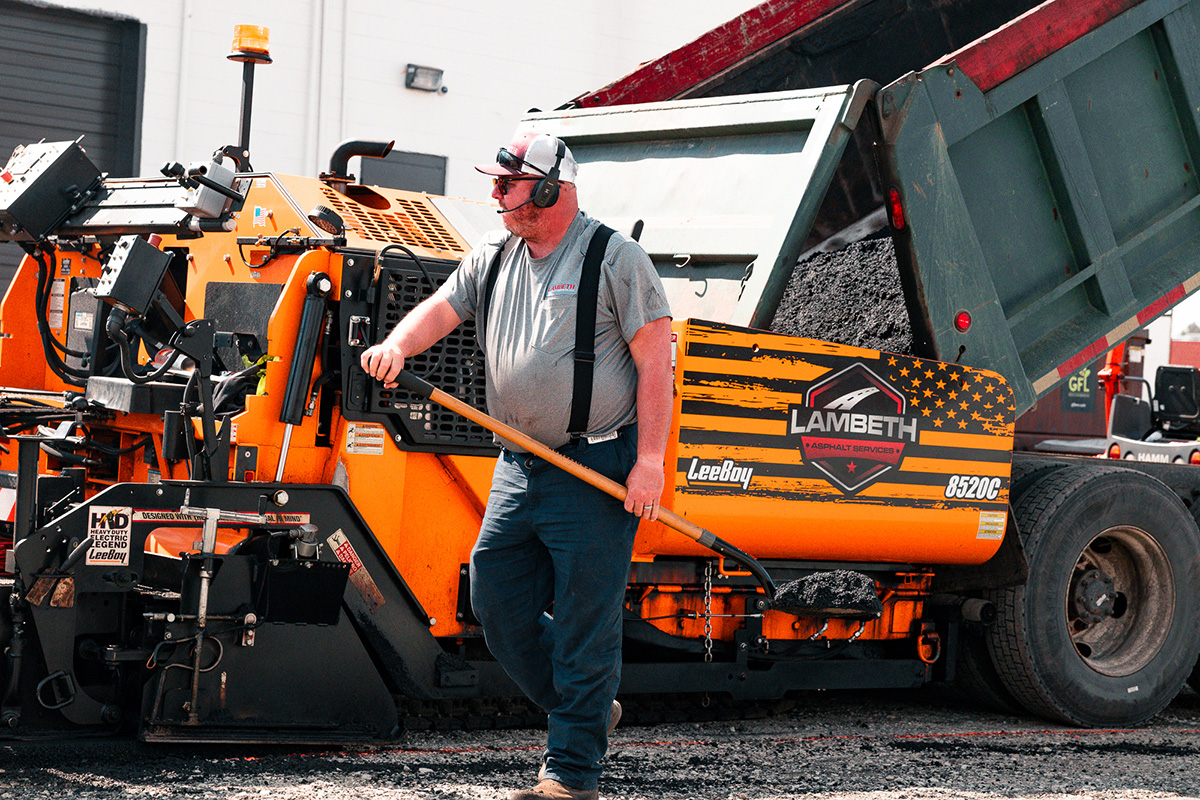 Lambeth Asphalt crew member working alongside a LeeBoy 8520C asphalt paver during full-depth parking lot replacement at NAI Piedmont Triad on Wendover Avenue, Greensboro NC