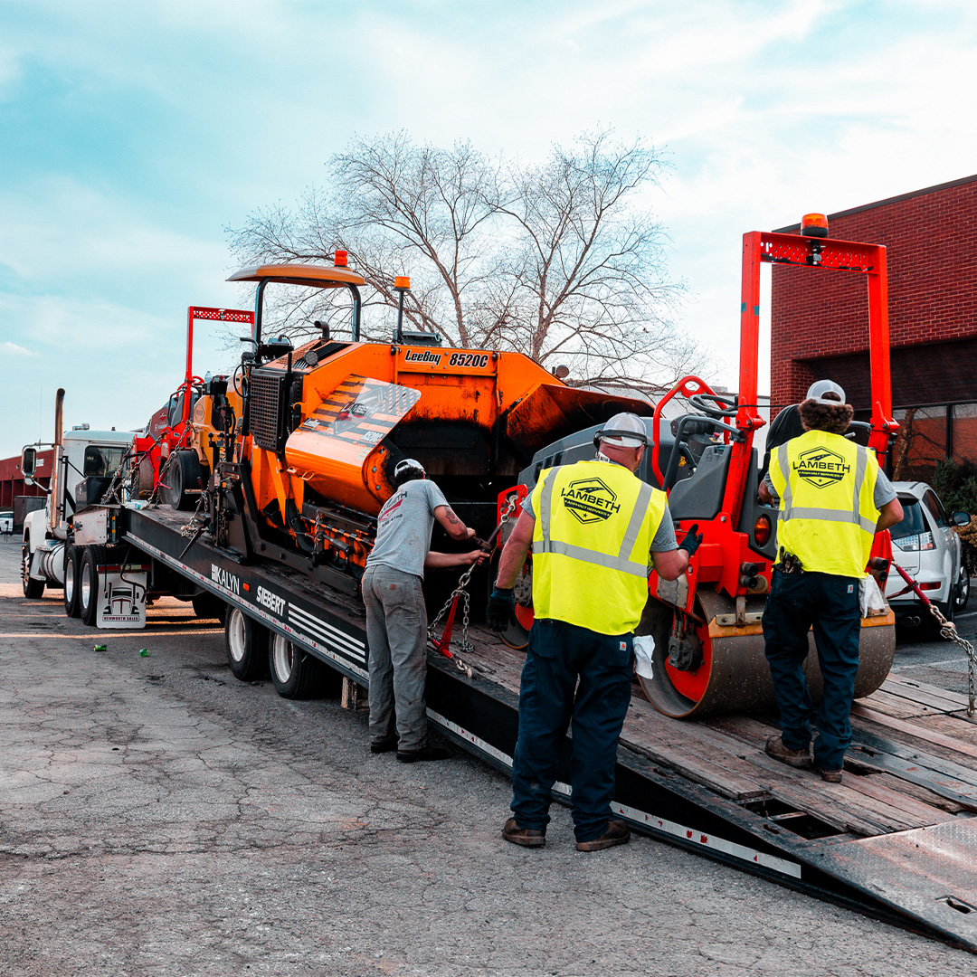 Lambeth Asphalt crew securing a LeeBoy 8520C paver and HAMM roller to a flatbed trailer before mobilizing to the NAI Piedmont Triad job site on Wendover Avenue, Greensboro NC