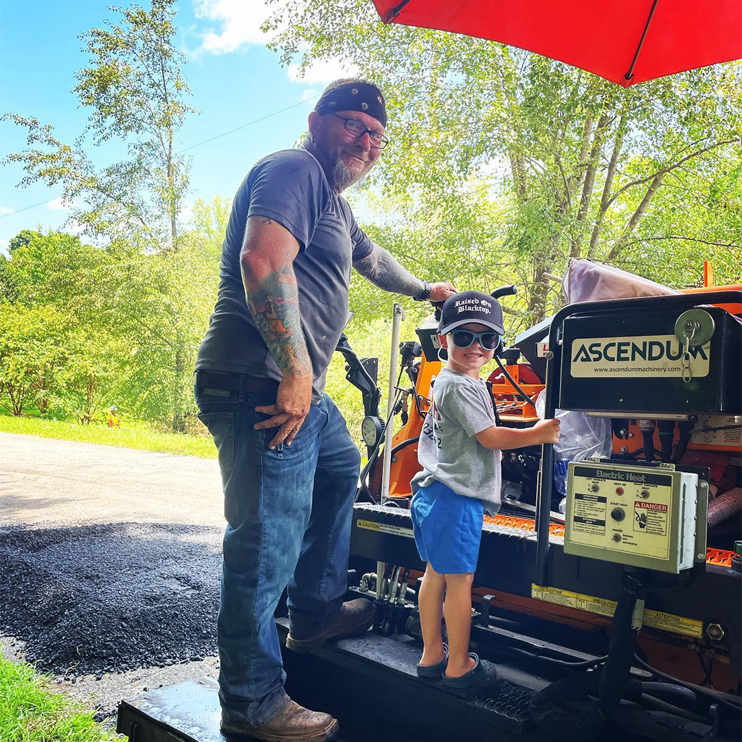 Lambeth Asphalt crew member and young child posing together on an Ascendum asphalt paver at a paving job site