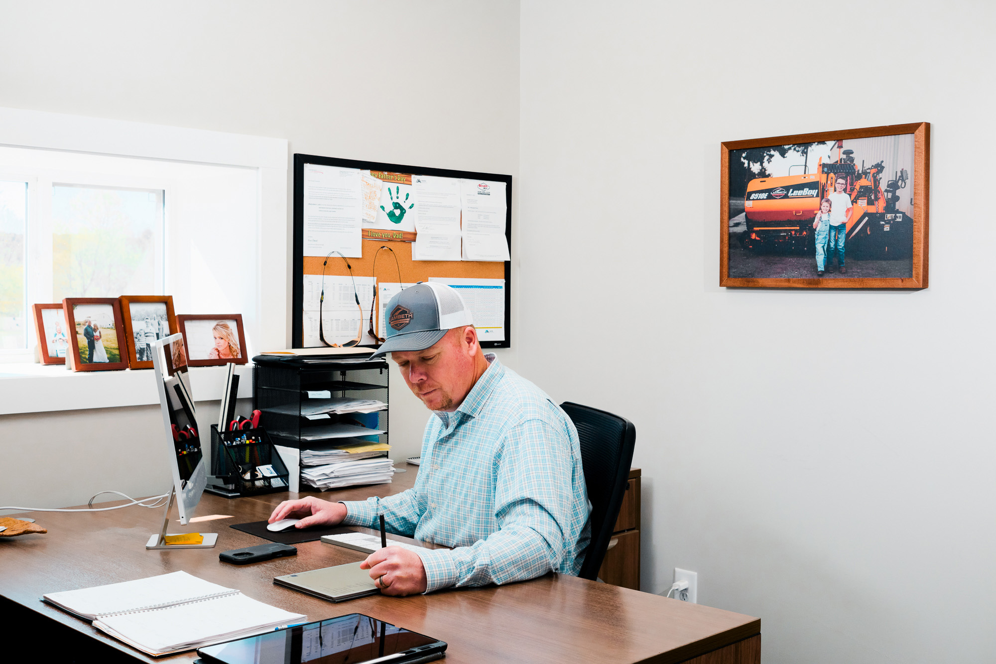 Man in a light blue checkered shirt and gray cap working at a desk with a computer, papers, and a tablet in a bright office with framed photos and a bulletin board.