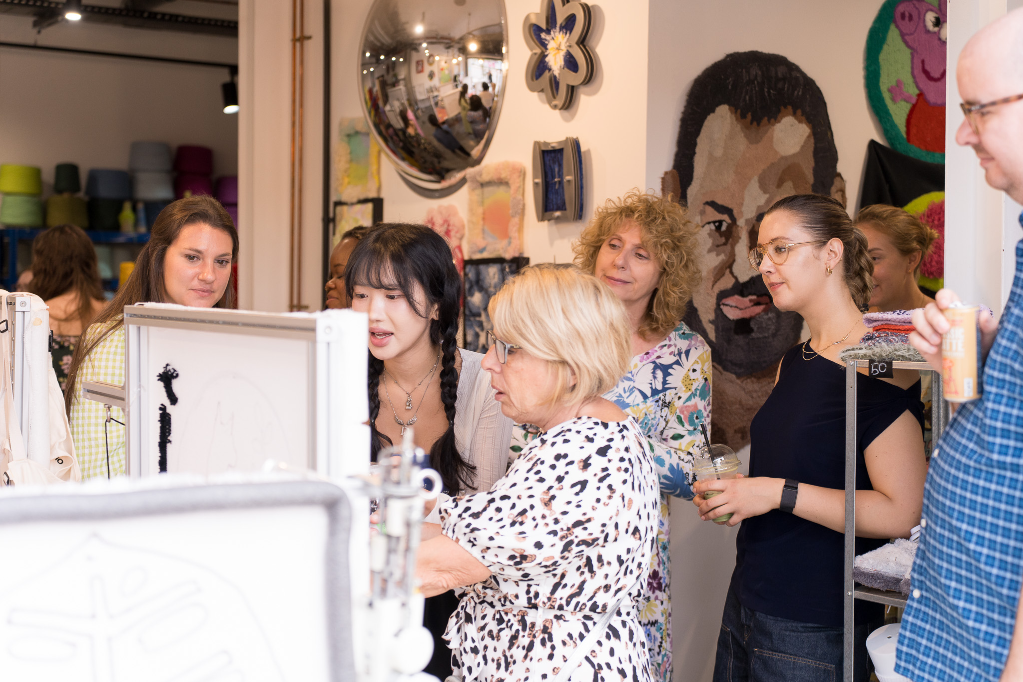 A diverse group of people observing and discussing artwork displayed on frames in a gallery setting with textile art and a large mosaic portrait in the background.
