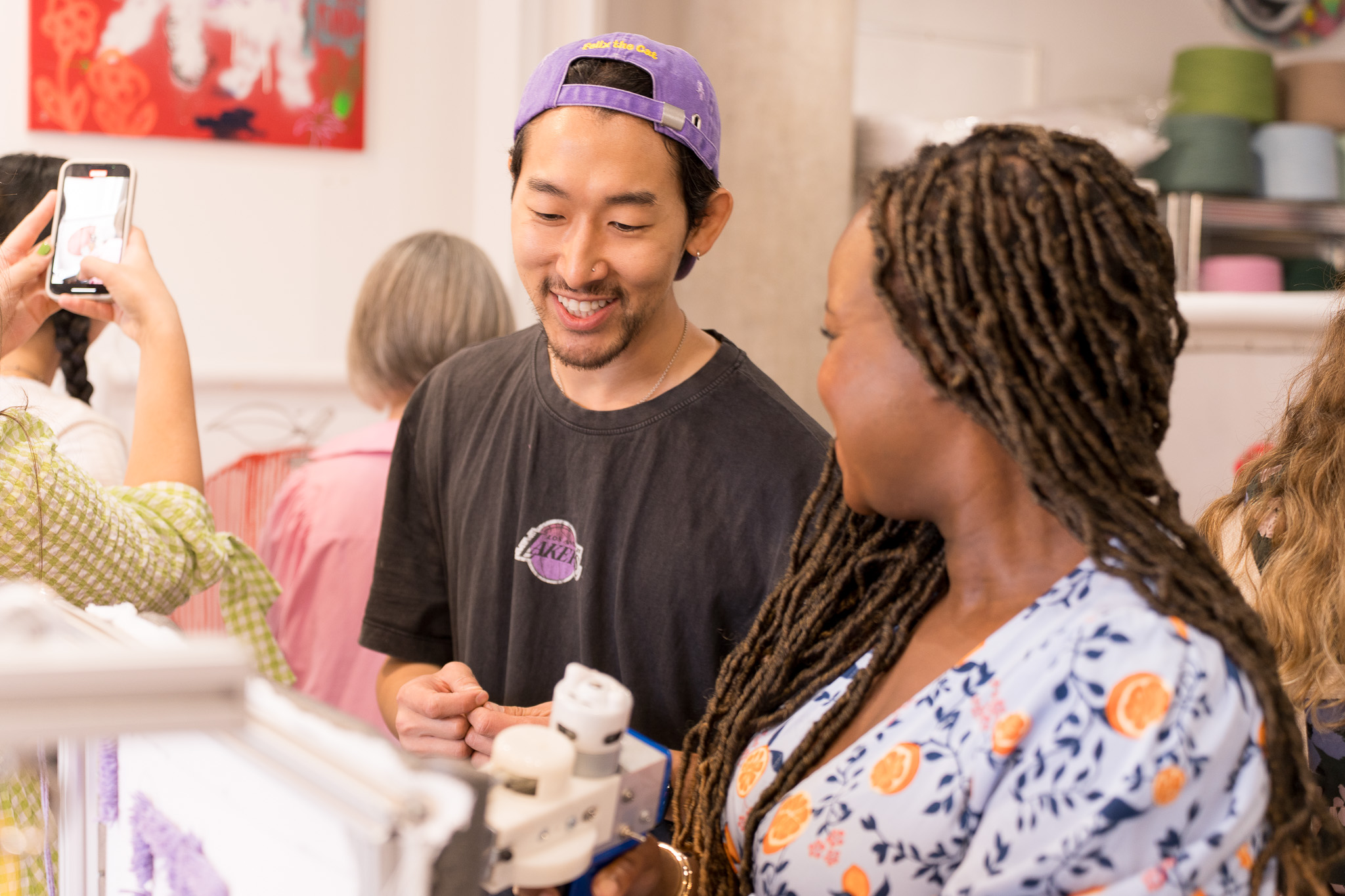 Two people smiling and interacting, with one wearing a purple cap and black t-shirt and the other with braided hair in a patterned blouse.