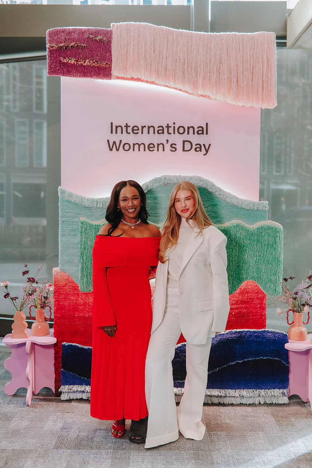 Two women standing and smiling in front of a colorful textured backdrop with 'International Women’s Day' text above.