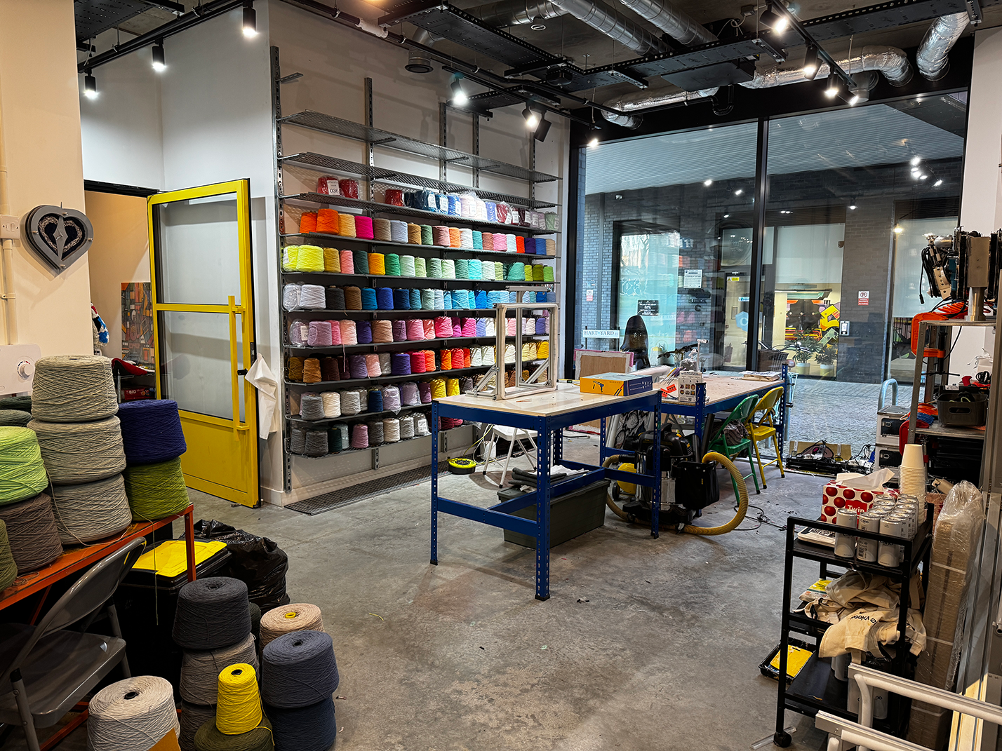 Interior of a craft workshop with shelves of colorful yarn spools, work tables, and large windows overlooking an outdoor area.