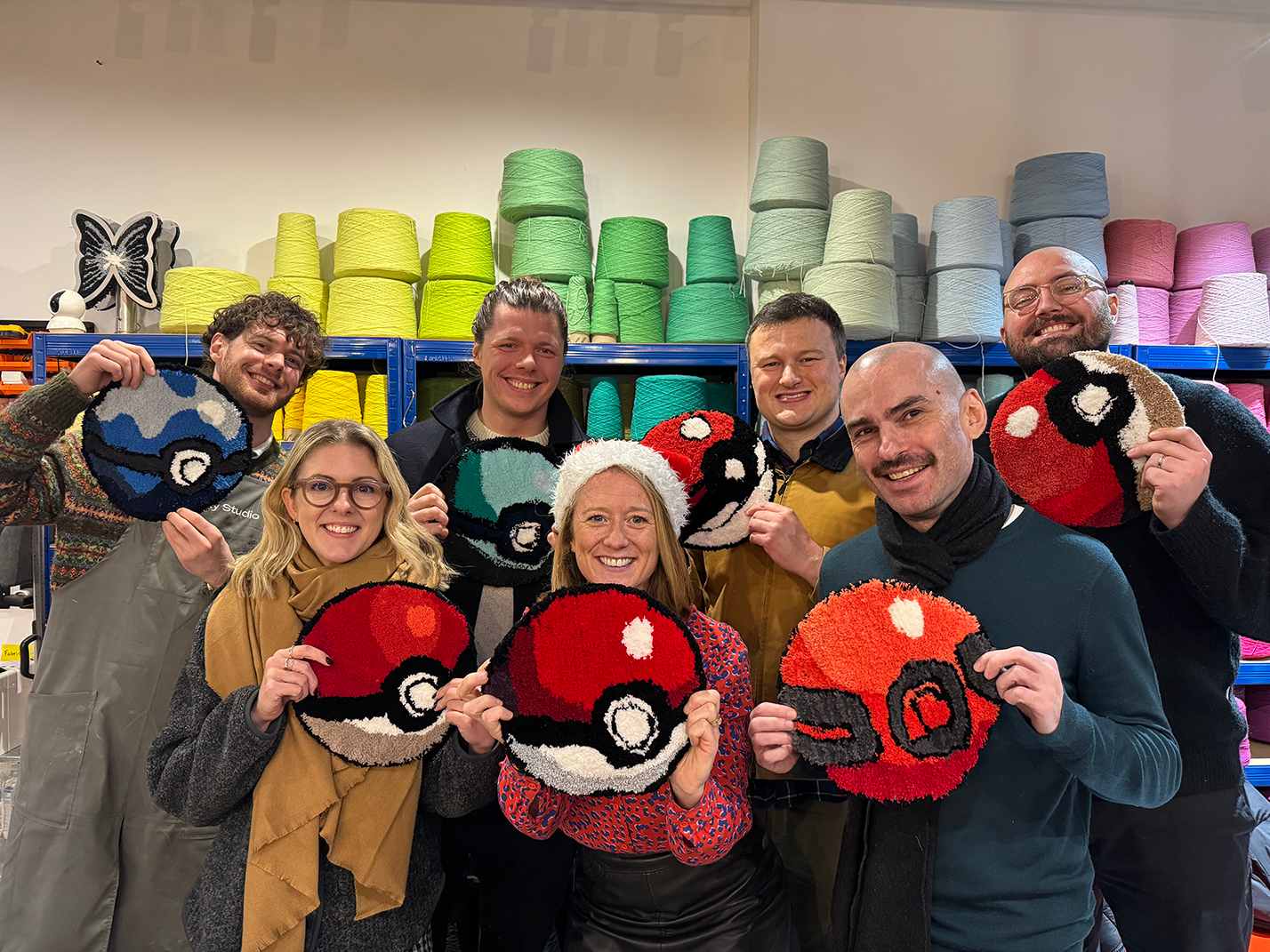 Group of seven people smiling and holding round crochet art pieces resembling various Poké Balls in a colorful yarn studio.