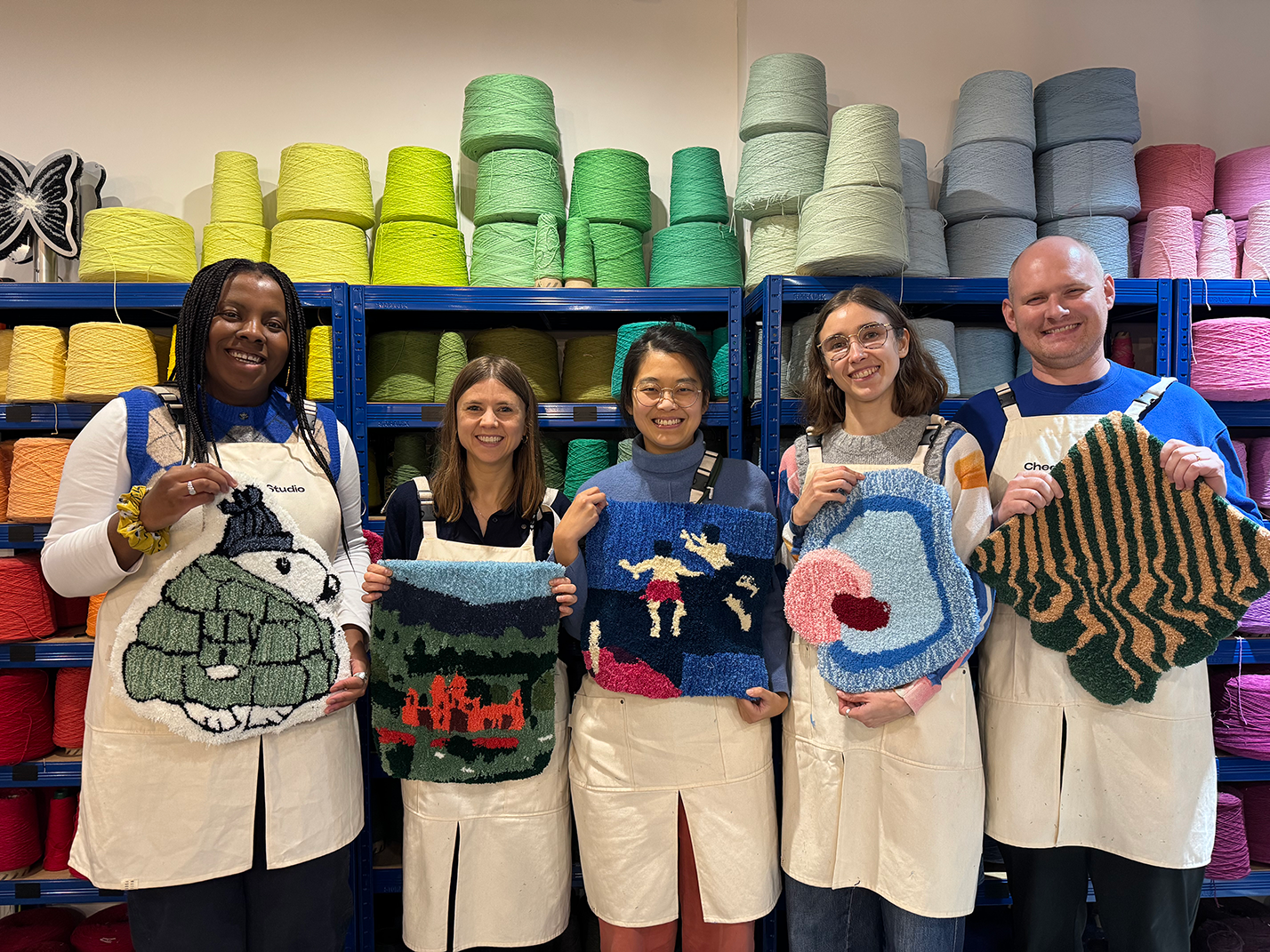 Five people standing in front of colorful yarn shelves, each holding a handmade tufted textile art piece with various designs.