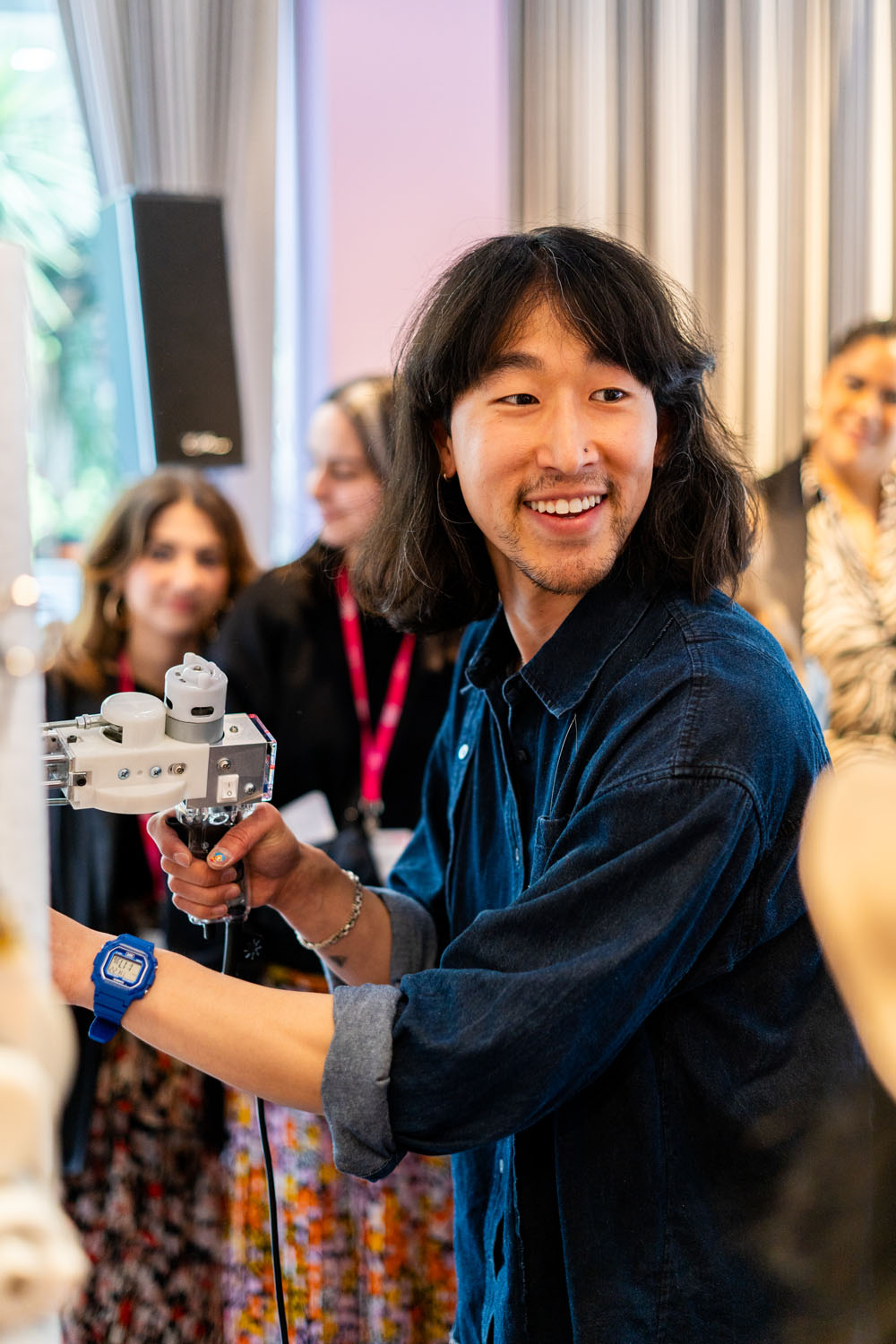 Young man in denim shirt smiling while holding and operating a robotic arm, with blurred people watching in the background.