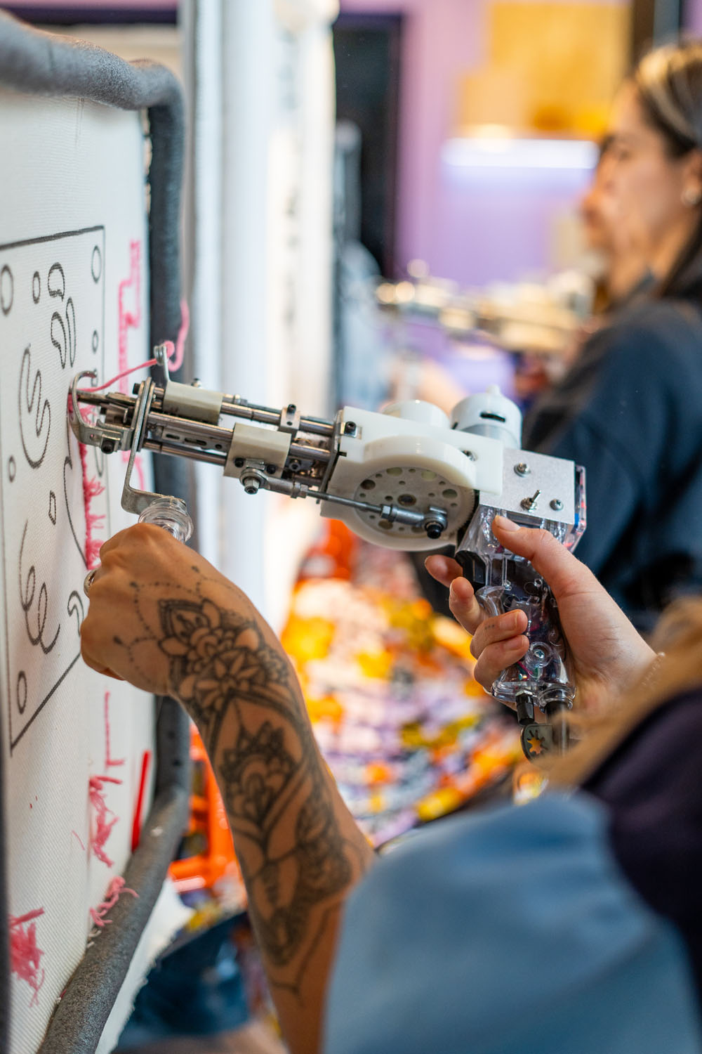 Person with tattooed arm using a mechanical embroidery machine on fabric stretched in a frame.