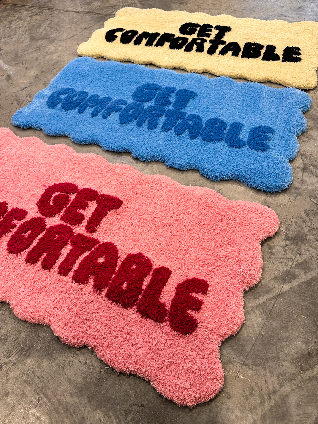 Three colorful shaggy rugs on a concrete floor with scalloped edges, each reading 'GET COMFORTABLE' in bold text; rugs are yellow with black text, blue with darker blue text, and pink with red text.