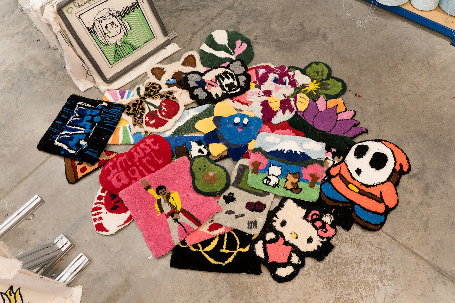 A collection of colorful, handcrafted rugs featuring various cartoon characters and designs scattered on a concrete floor in an art studio.