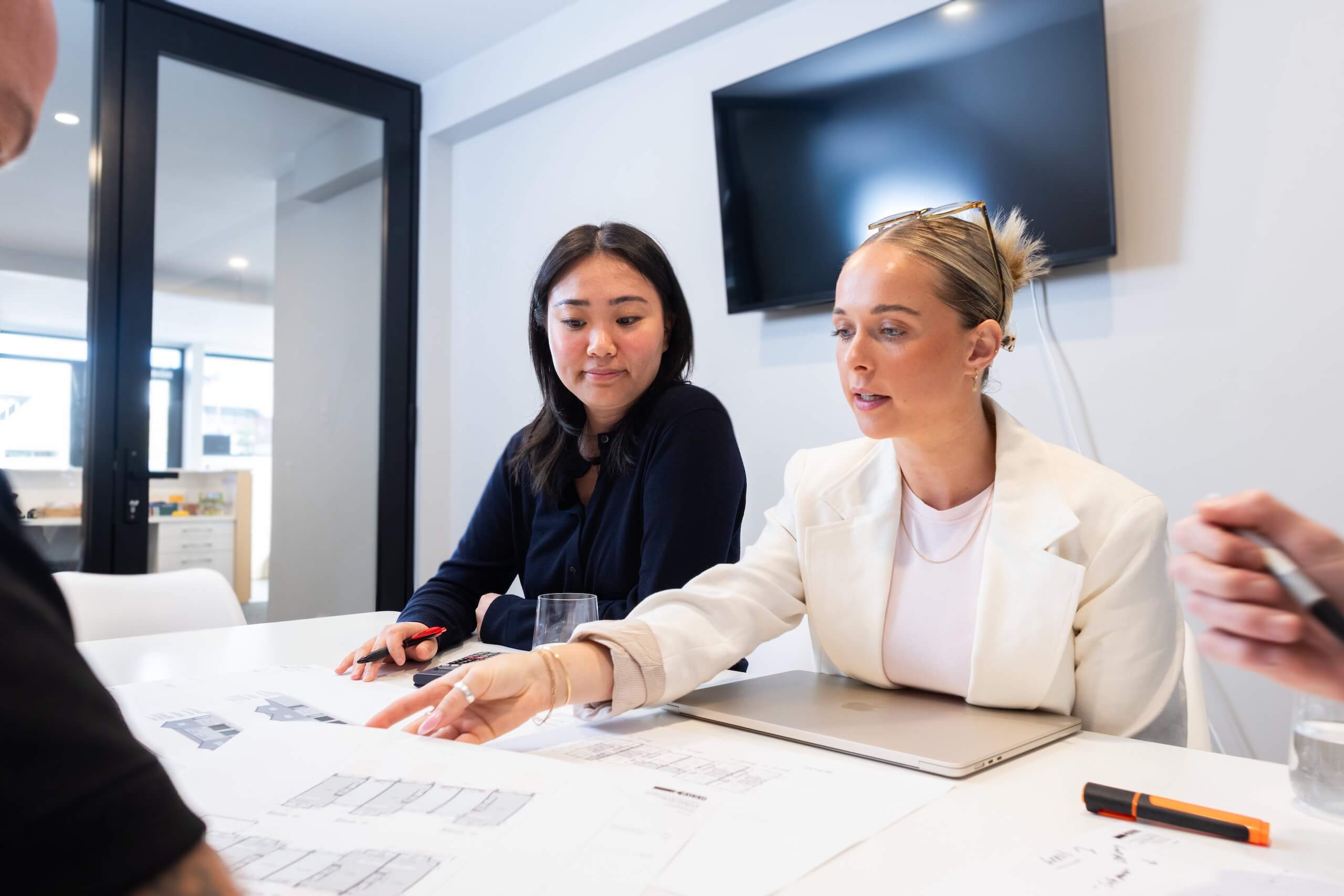 Two women discussing architectural floor plans at a table with a laptop and stationery in an office.