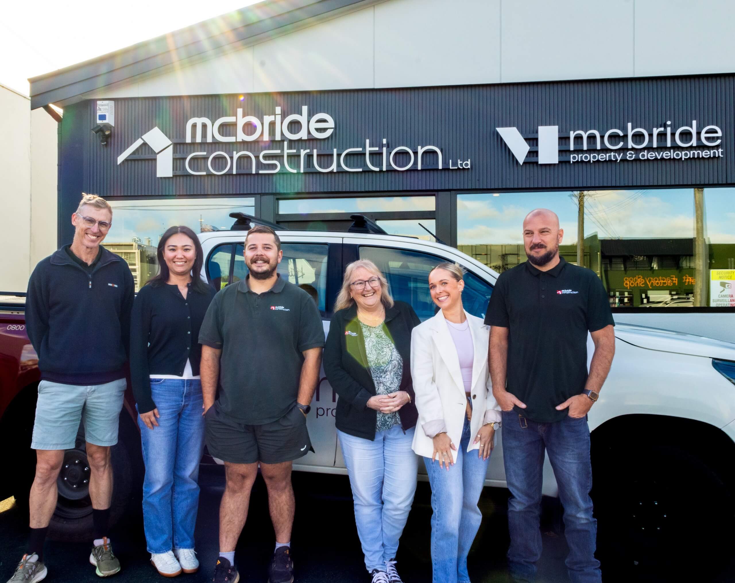 Six smiling McBride Construction and Property & Development employees standing in front of a company vehicle and building.
