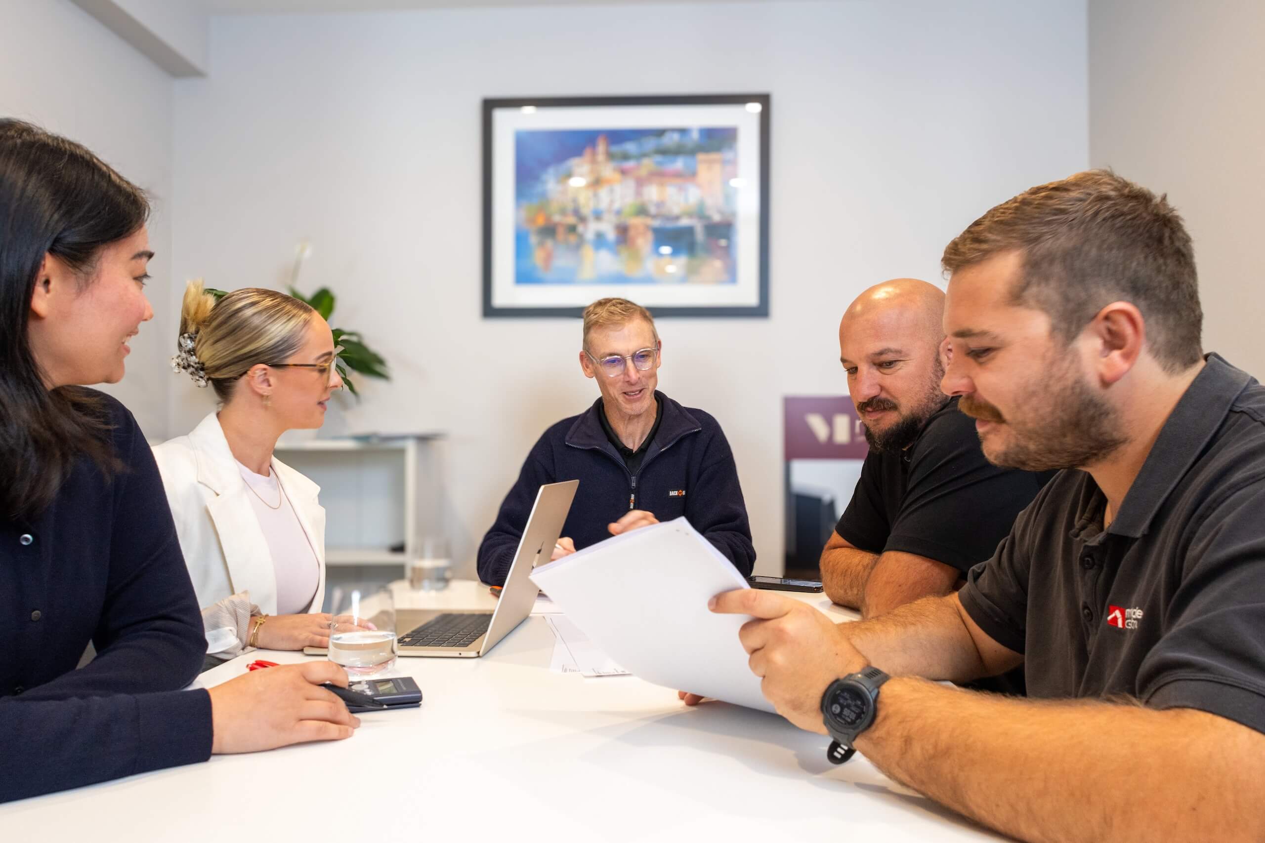 Five colleagues around a table in a meeting room reviewing documents and using a laptop.