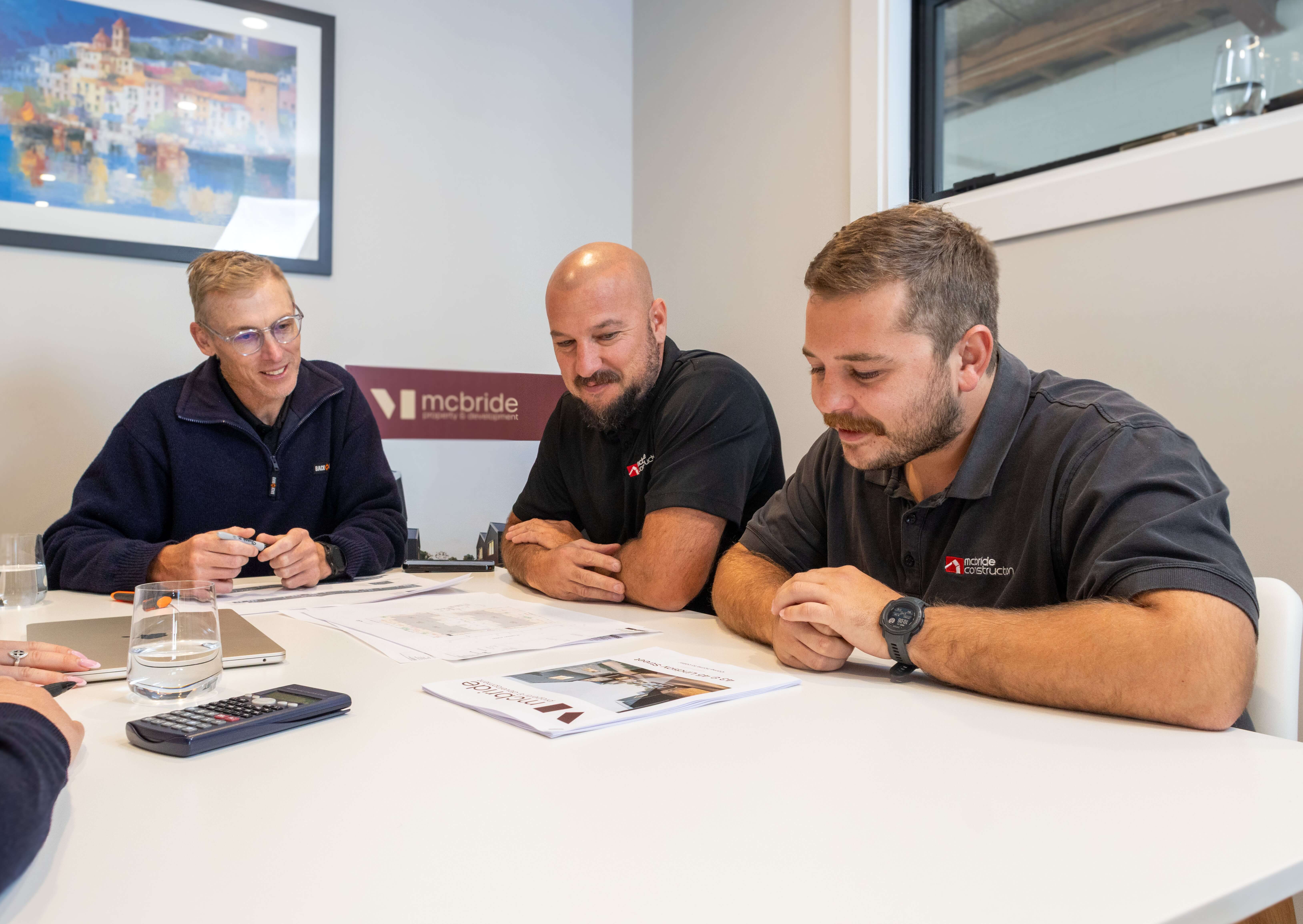 Three men sitting around a table reviewing documents and plans in an office with a McBride sign on the wall.