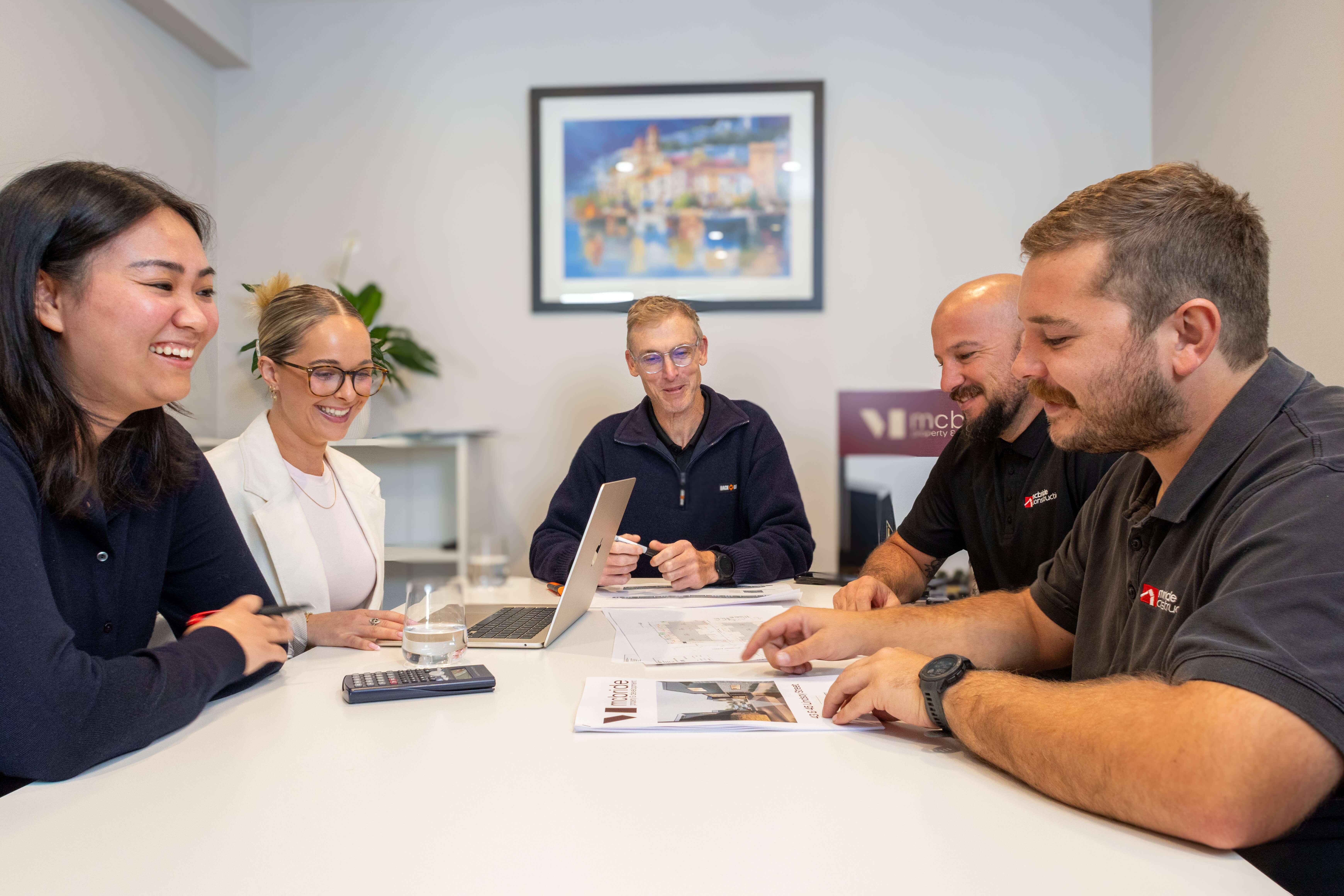 Five people sitting around a table in a meeting room, smiling and discussing documents and a laptop.