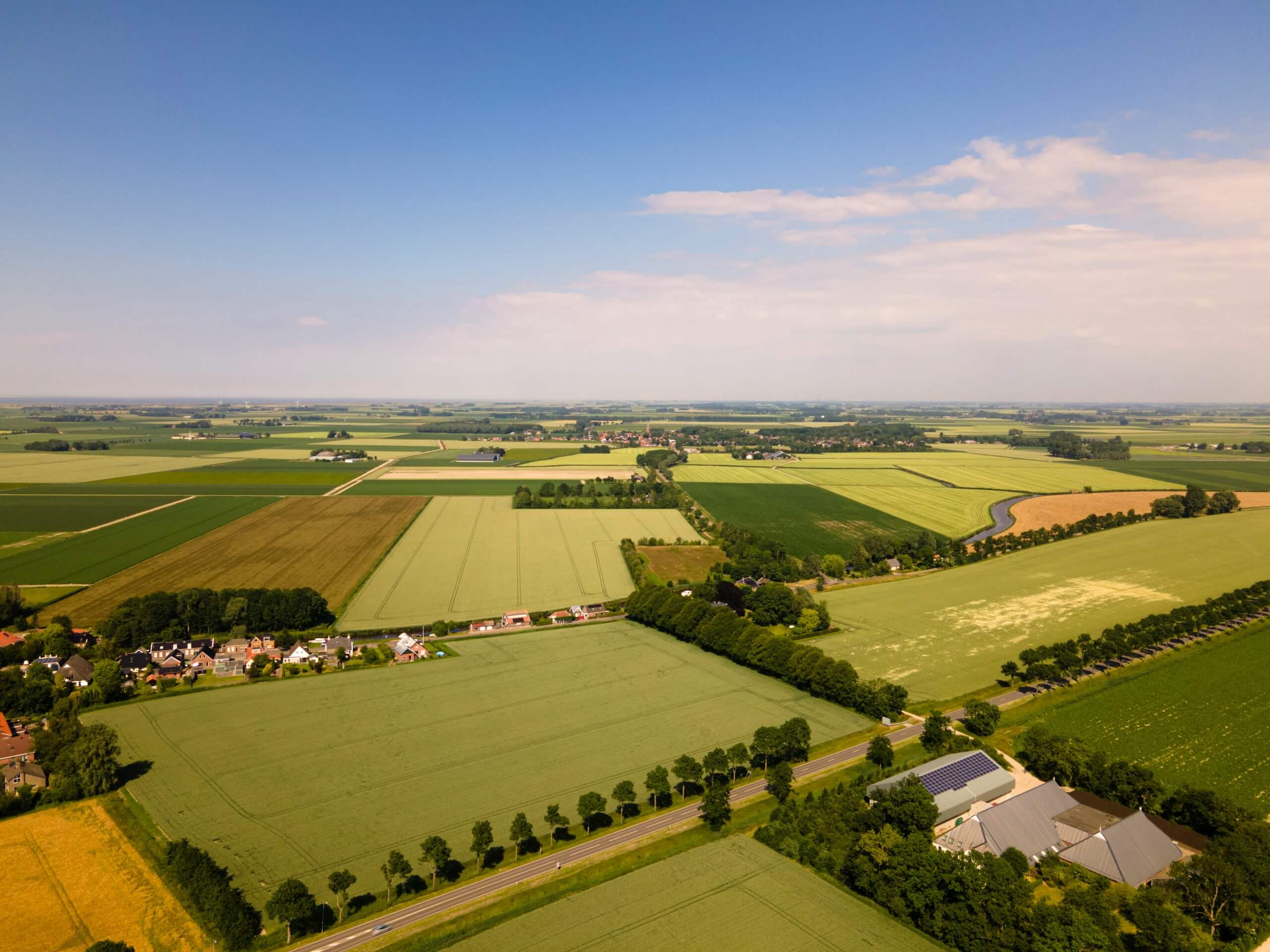 Aerial view of a rural landscape with cultivated fields, a small village, roads lined with trees, and farm buildings under a partly cloudy sky.