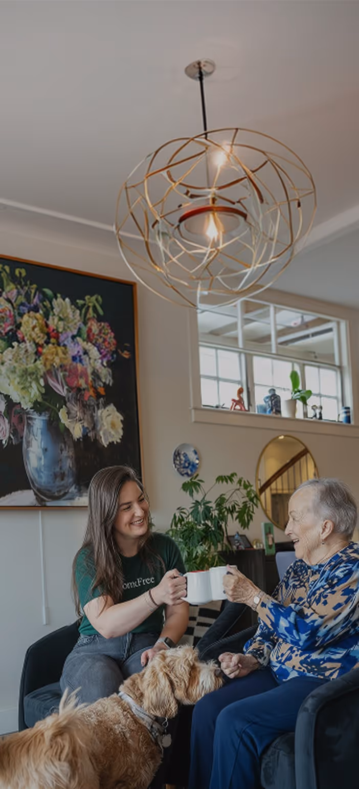 Young woman and elderly woman sitting, clinking white mugs, with a dog nearby in a cozy living room.