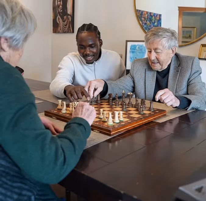 Two elderly people and a young man playing chess together at a dining table in a cozy room.