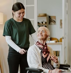 A young woman in green stands behind an elderly woman in a wheelchair, both smiling indoors.