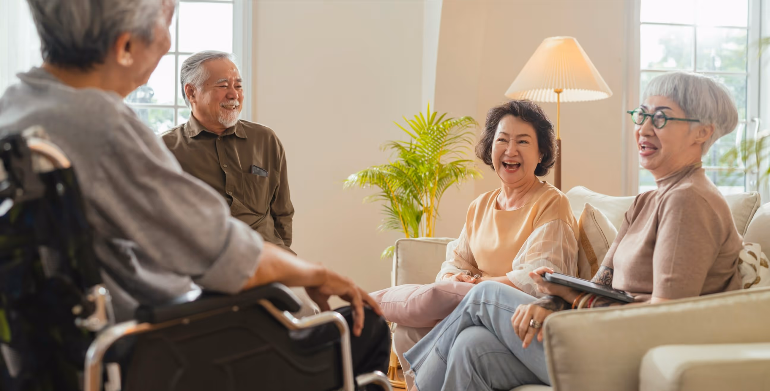 Four elderly Asian adults smiling and chatting in a cozy living room, one seated in a wheelchair.