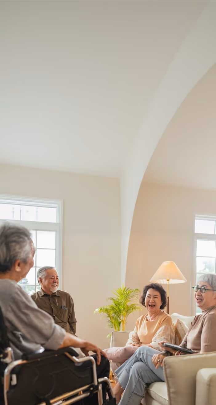 Four elderly Asian adults smiling and talking in a bright, cozy living room.