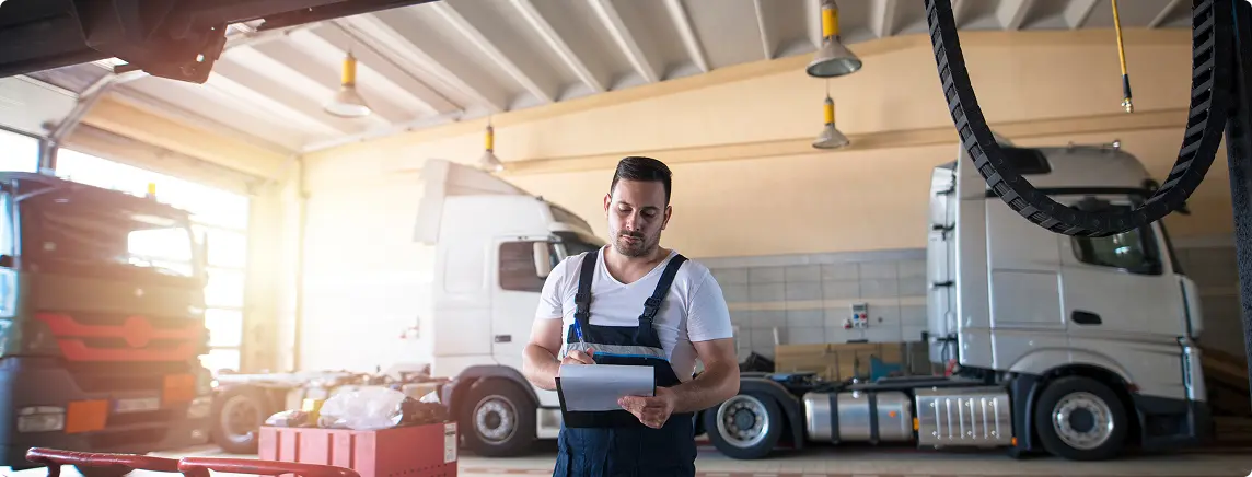 Mechanic in overalls writing on clipboard in garage with two large trucks in the background.