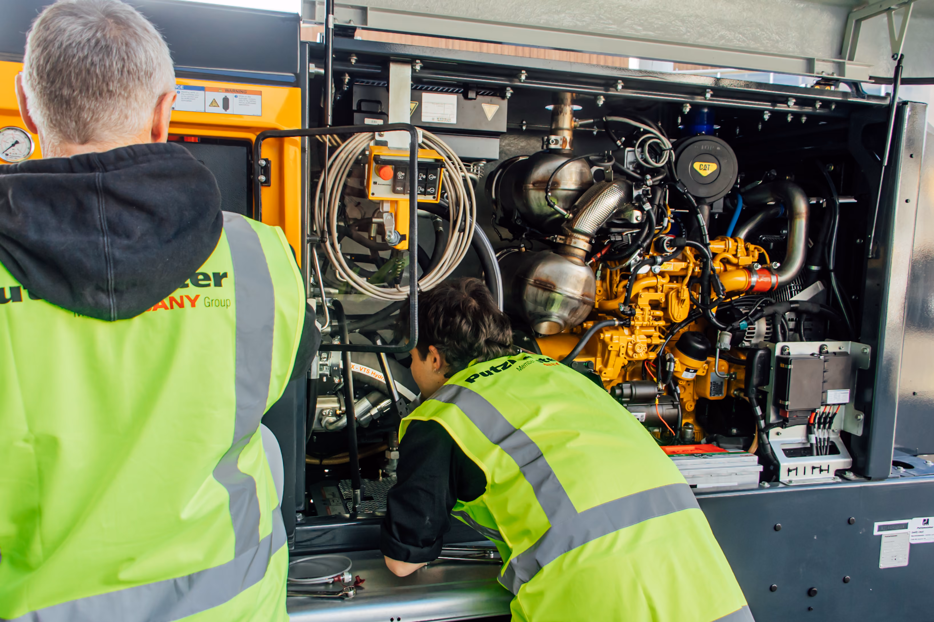 Two workers in yellow safety vests inspecting the engine of industrial machinery.