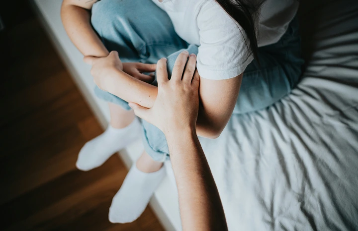 Close-up of one person sitting on a bed with legs crossed while another person gently holds their arm and hand.