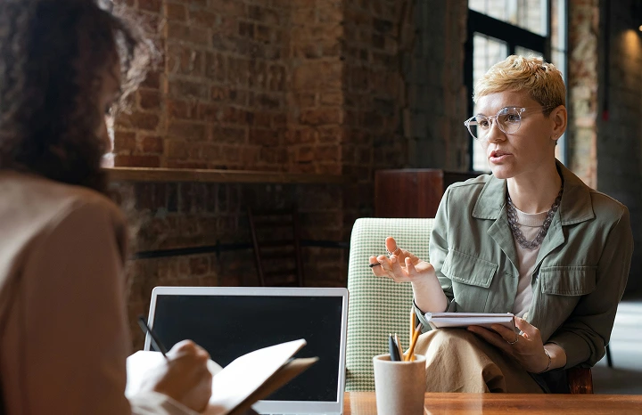 Two women engaged in a conversation during a meeting in a cozy office with brick walls; one is speaking while holding a tablet, the other is taking notes.