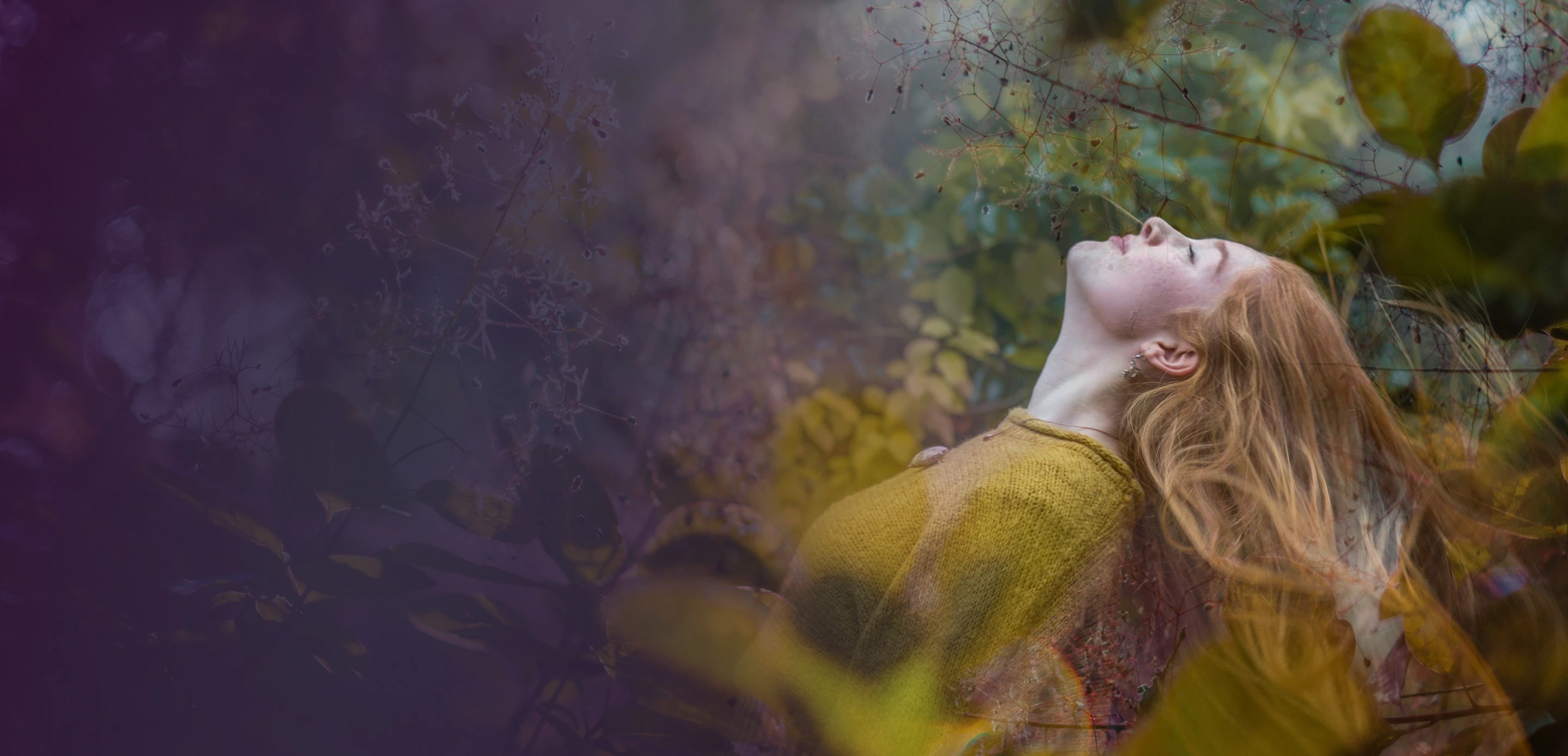 Woman with red hair and mustard sweater leaning back with closed eyes surrounded by blurred foliage.
