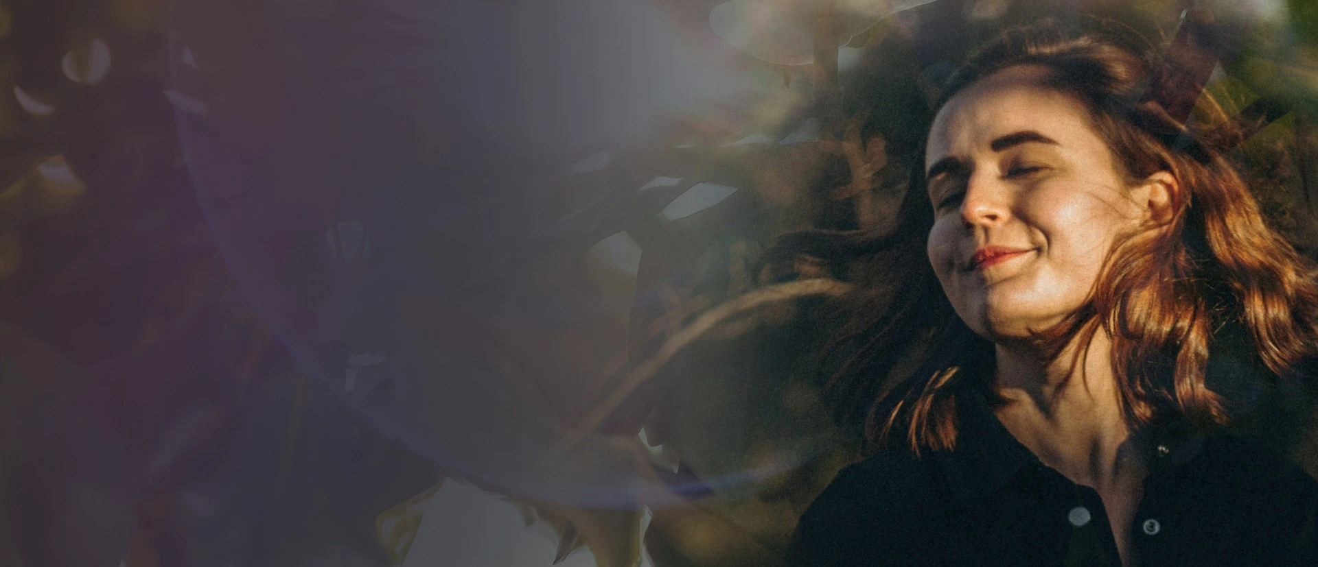 Woman with auburn hair smiling contentedly with eyes closed, surrounded by blurred foliage.