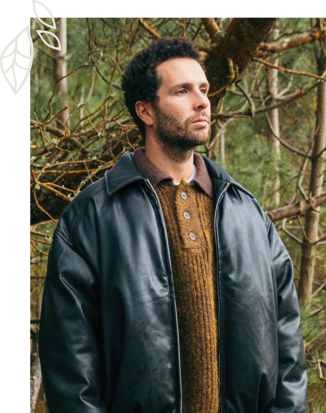 Man with curly hair and beard wearing a black leather jacket and brown knit sweater standing in a forest.