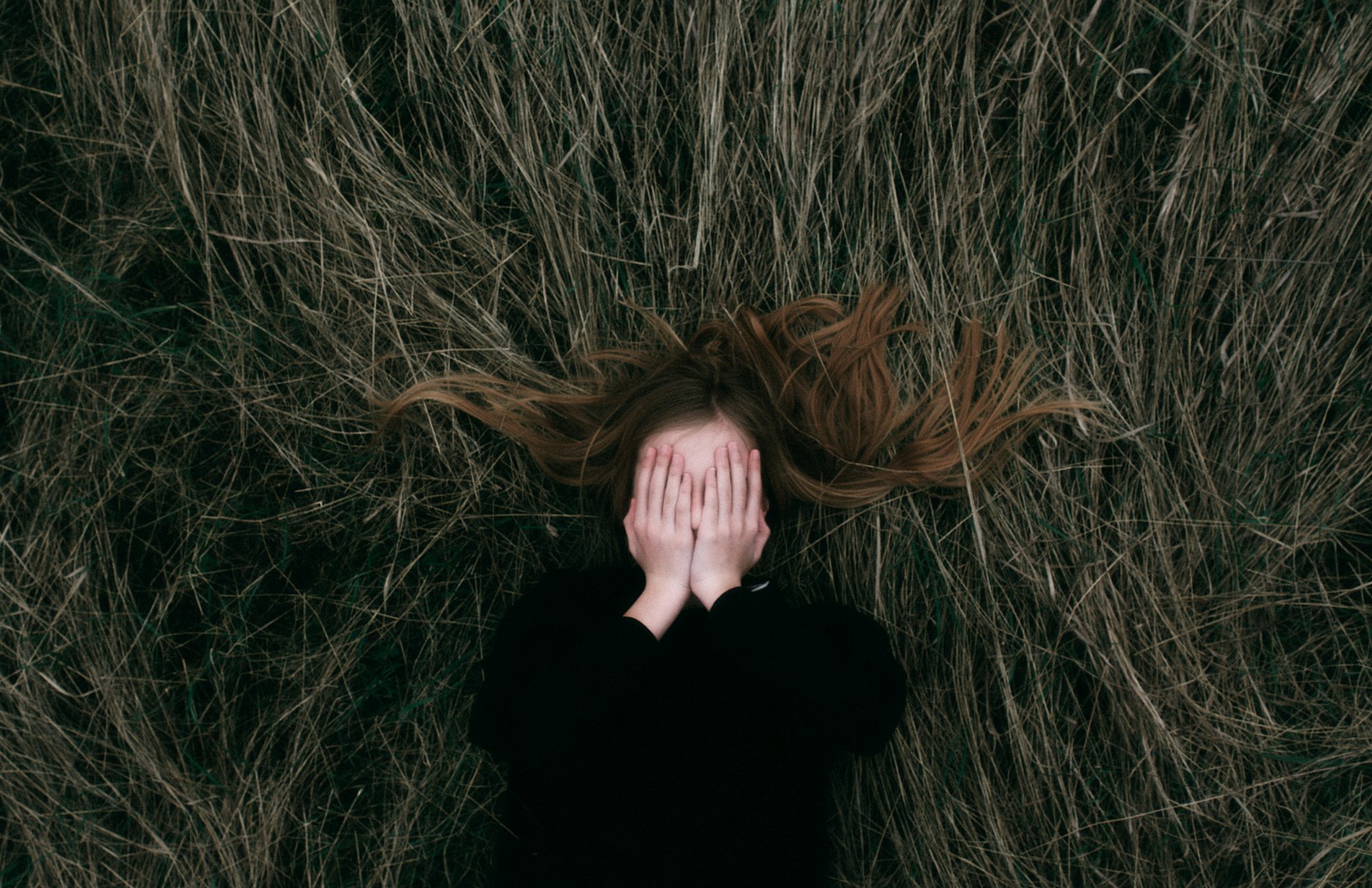 Person lying on dry grass with hands covering their face and hair spread around their head.
