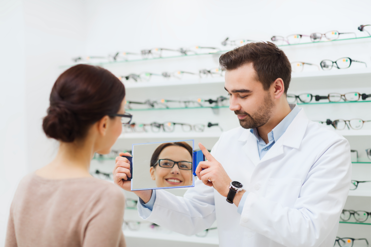 Optician in white coat showing a woman her glasses reflection in a mirror at an eyewear store.