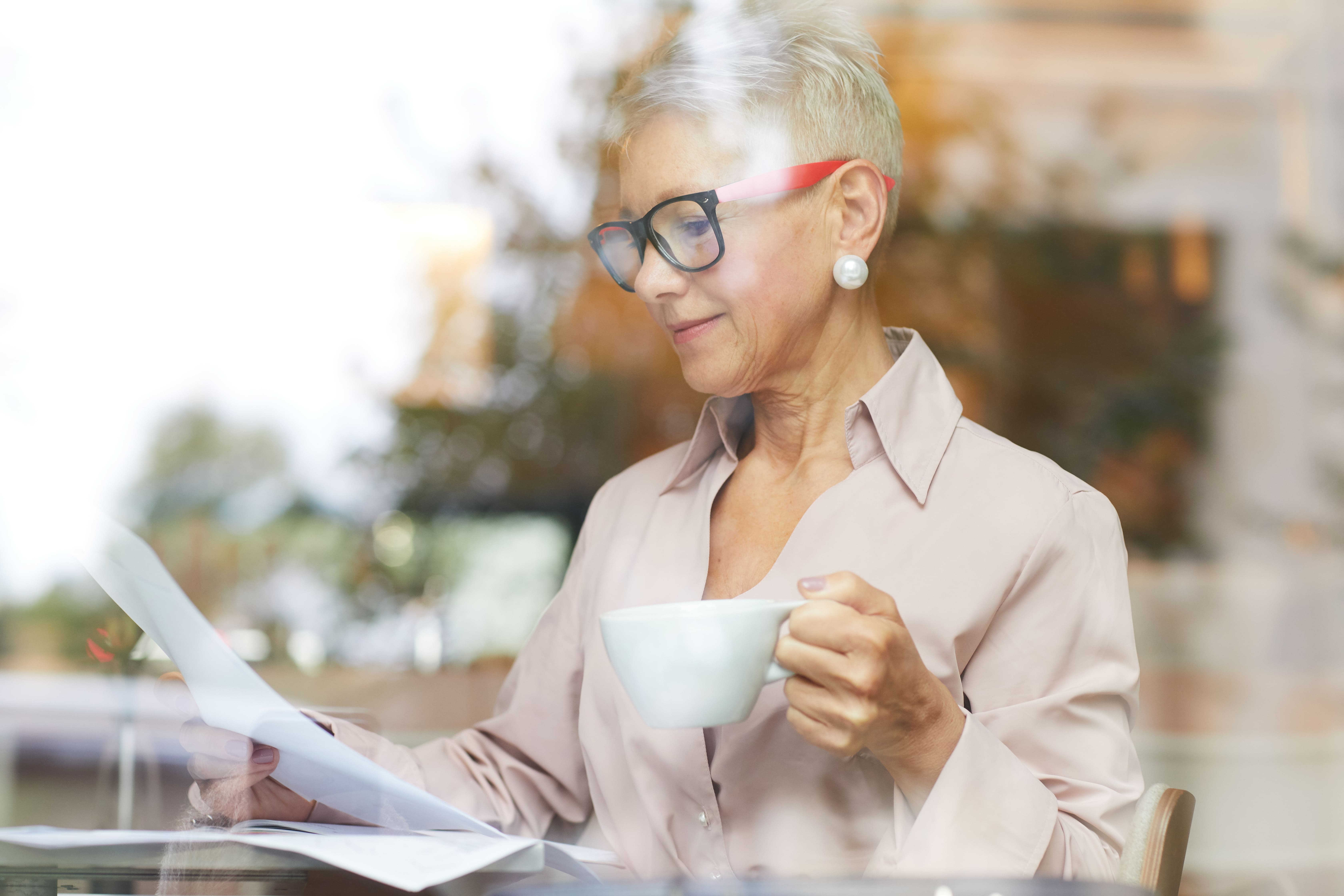 Mature woman with short hair and glasses reading documents while holding a white cup indoors.