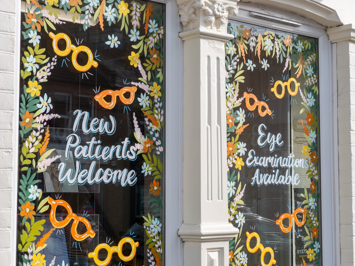 Store window with floral designs and signs reading New Patients Welcome and Eye Examinations Available.