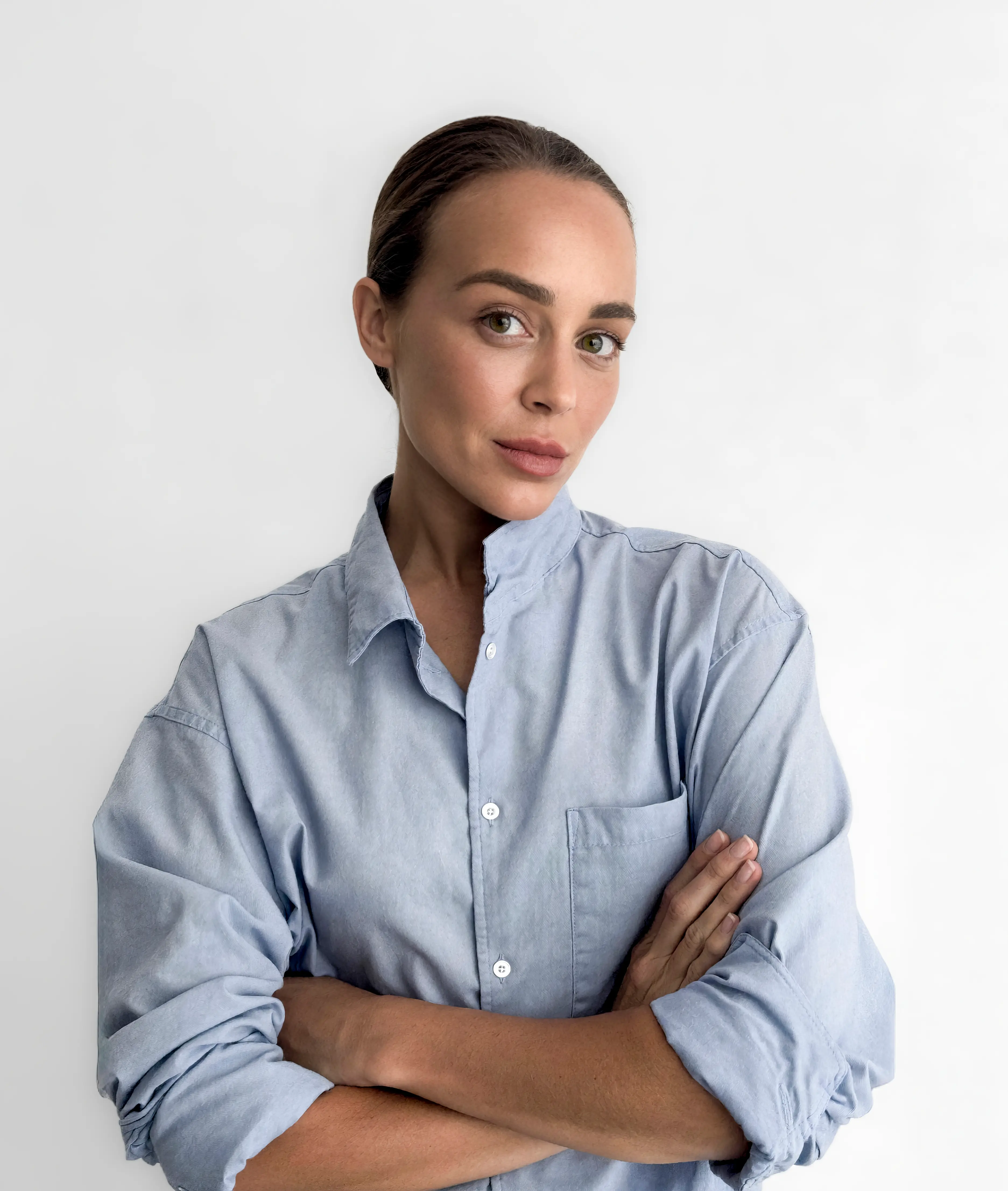 Young woman with slicked-back hair wearing a light blue button-up shirt with sleeves rolled up, standing with arms crossed against a plain white background.