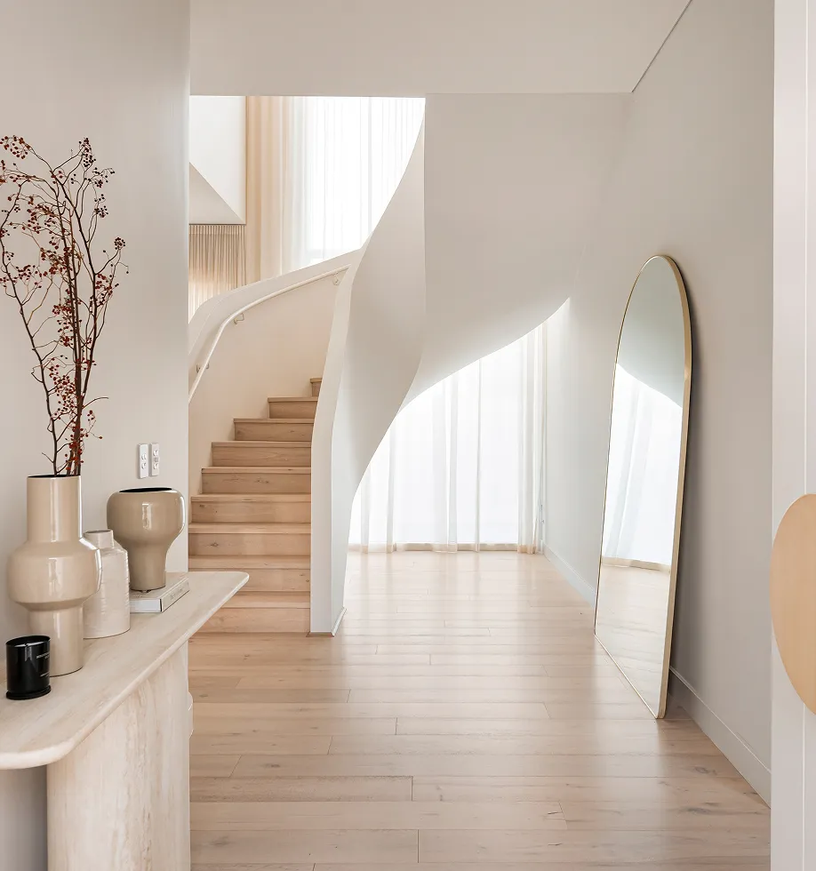 Minimalist interior with light wood flooring, curved white staircase, tall floor mirror, and a console table with ceramic vases and dried branches.