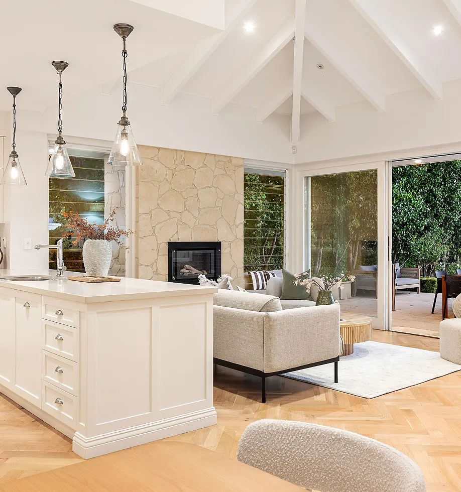 Bright open-plan living area with kitchen island, pendant lights, beige sofas, stone fireplace, and large sliding doors opening to a garden patio.