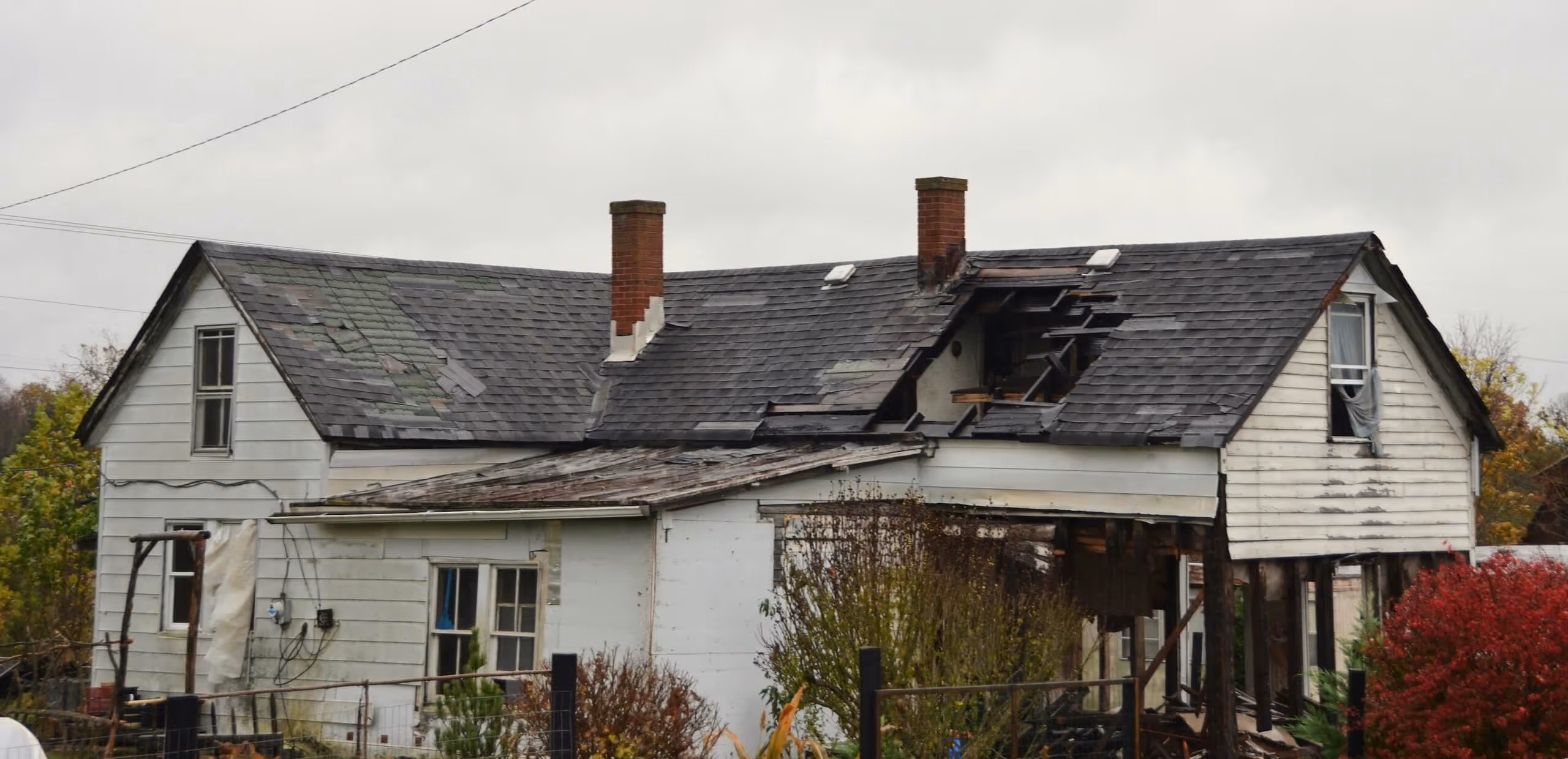Old white house with damaged roof and visible structural decay on a cloudy day.