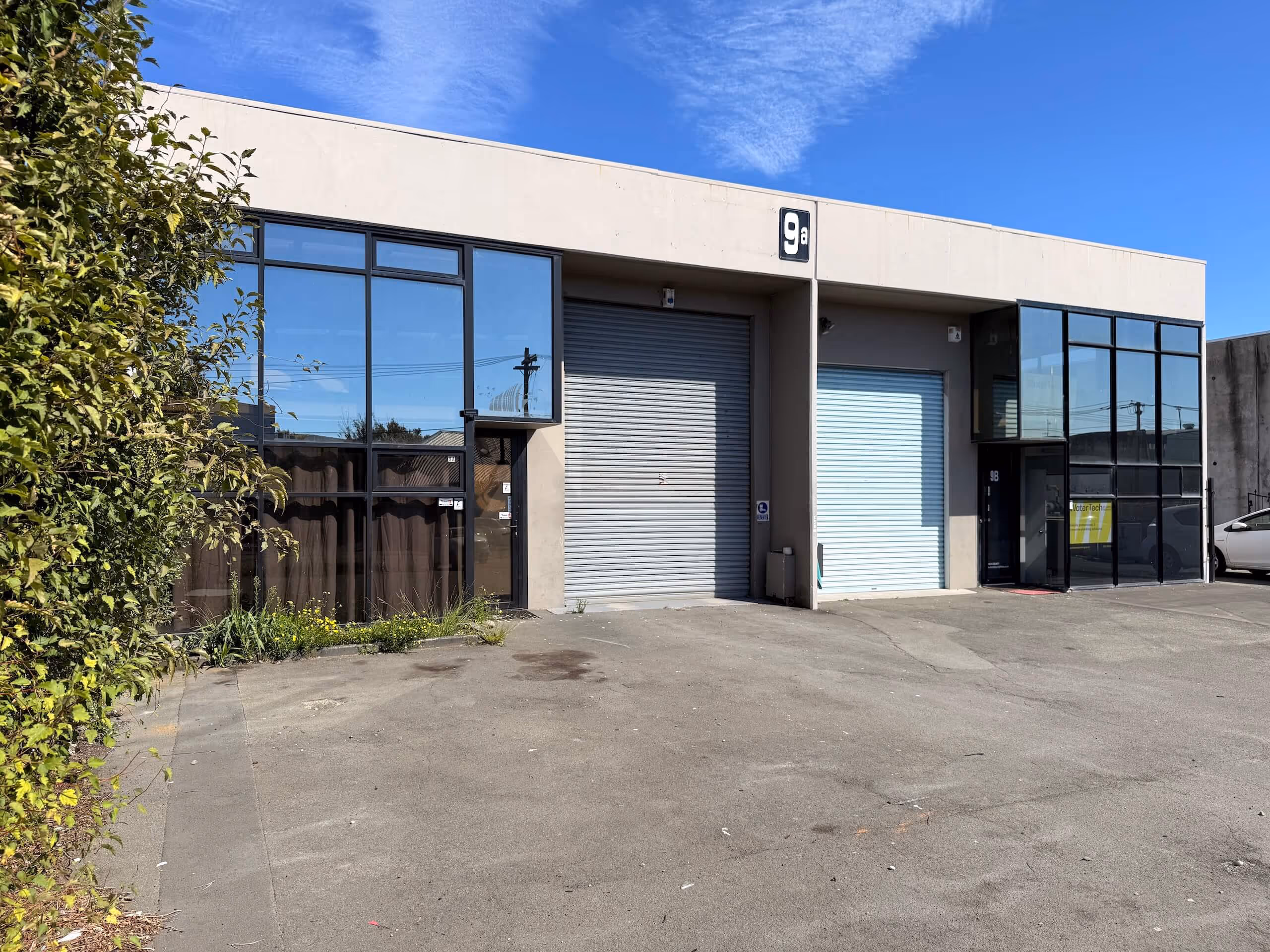 Two adjacent commercial units with large glass windows, roller shutter doors, and a concrete parking area under a clear blue sky.